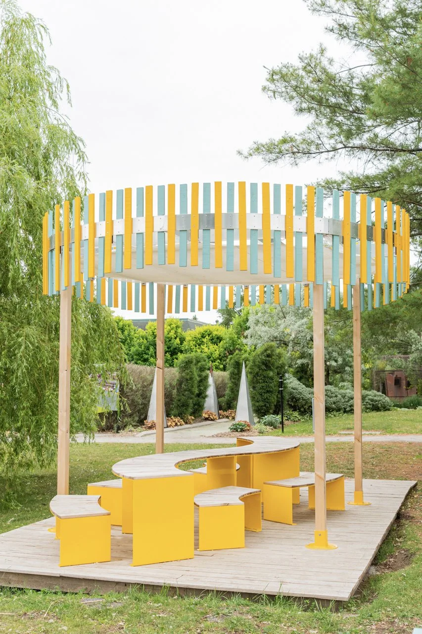 Structure de jeu colorée en jaune, bleu et blanc avec sièges intégrés, situ��e dans un parc avec des arbres verts et un chemin en arrière-plan.