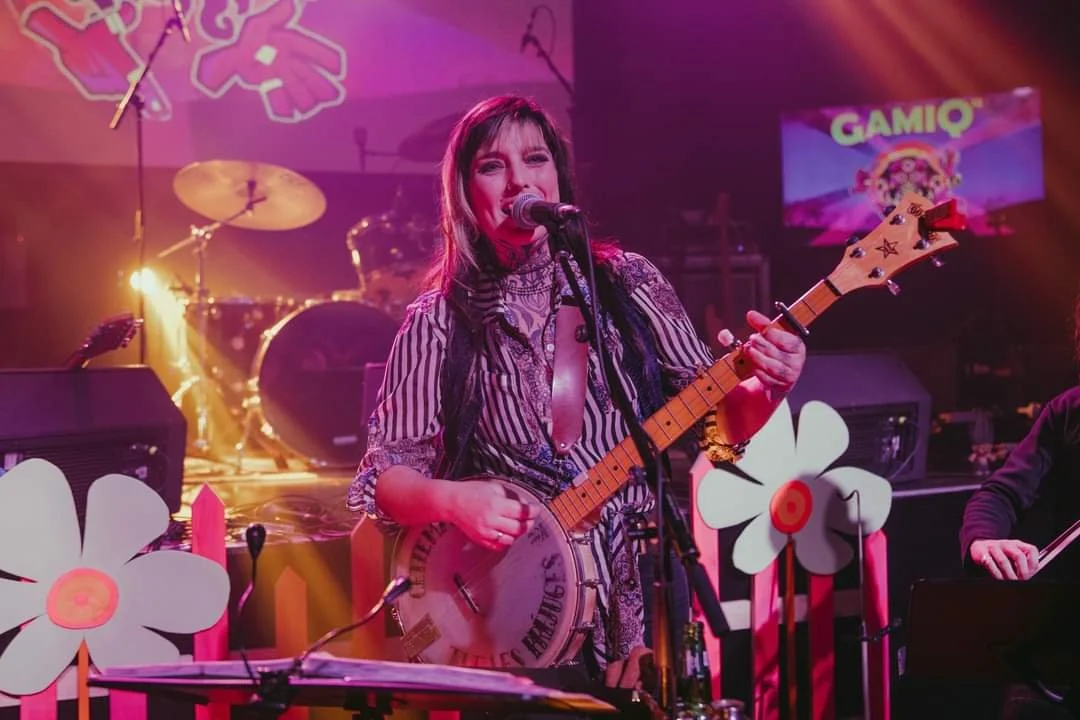 Une femme joue de la guitare et chante sur scène, entourée de décorations avec de grandes fleurs en papier et un fond coloré, lors d'un concert ou événement.