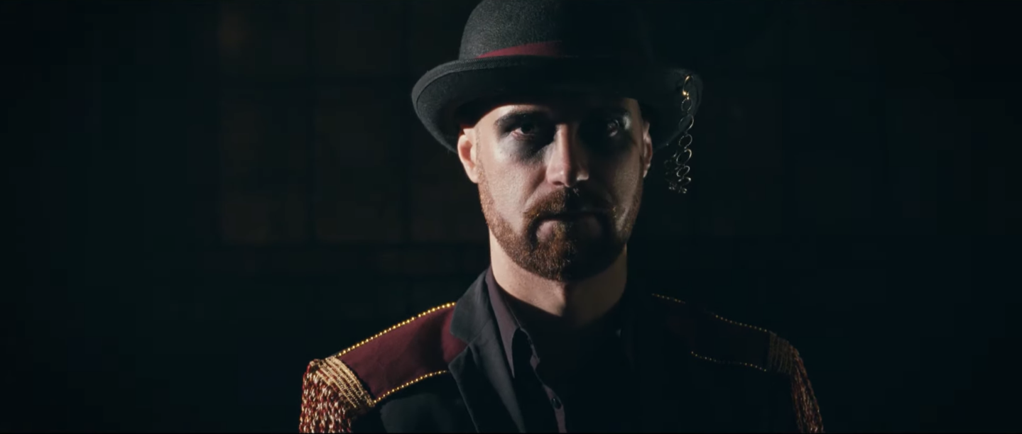 Un homme avec une barbe, portant un chapeau gris avec une chaîne, dans un éclairage sombre, portant une veste avec des épaulettes dorées.