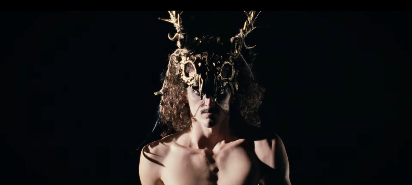 Une personne portant un masque ou une coiffe élaborée avec des éléments en forme de crâne et de cornes, dans un fond sombre.