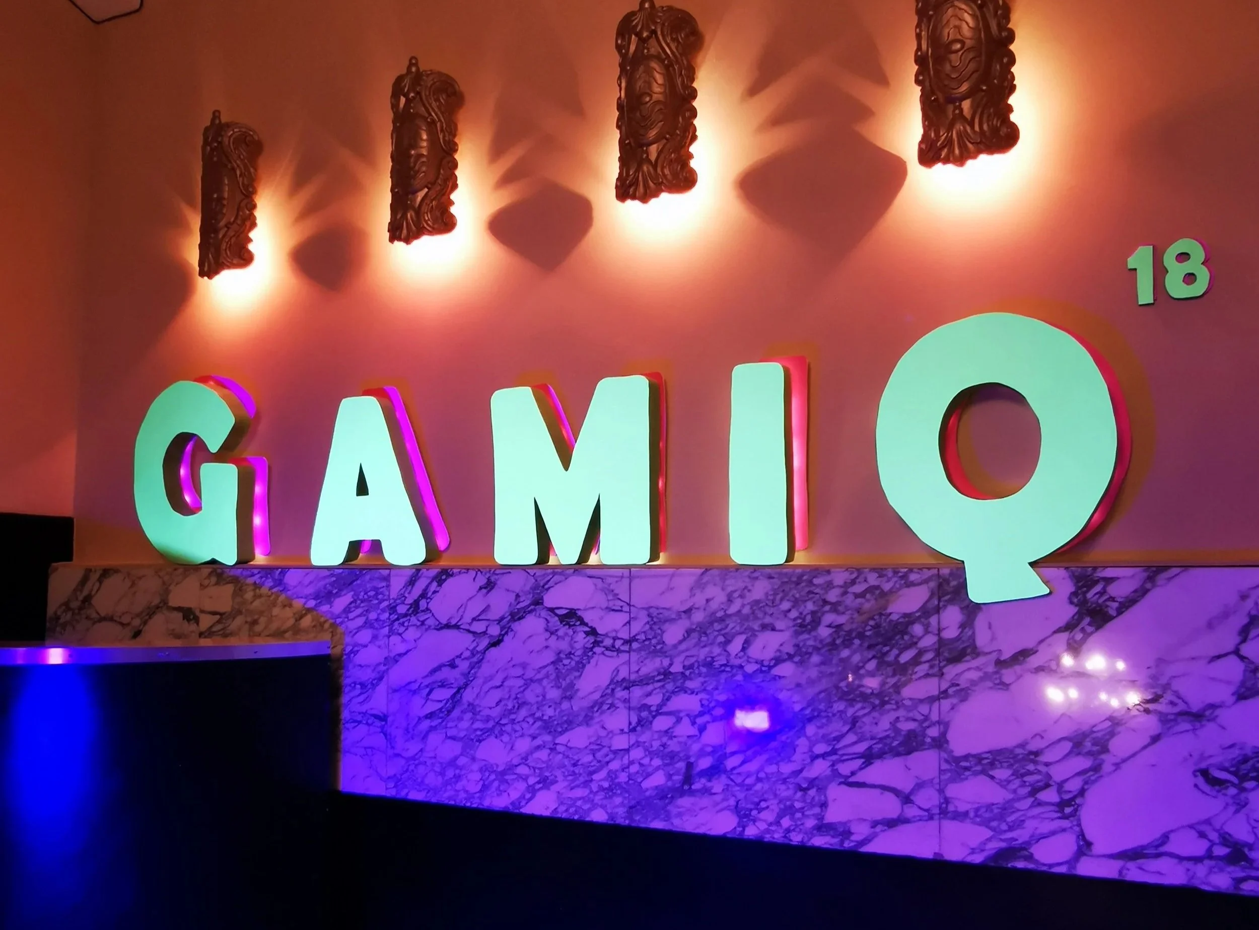 Lettres lumineuses formant le mot "GAMIC" avec décorations sculpturales suspendues au plafond, éclairées par un éclairage chaleureux sur un mur rose.