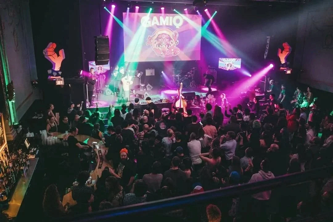 Scène de concert ou de spectacle avec une foule assise et debout devant une scène éclairée de lumières colorées, avec un écran géant affichant le mot 'GAMIO' et des illustrations, et de grandes mains en poing comme décor.