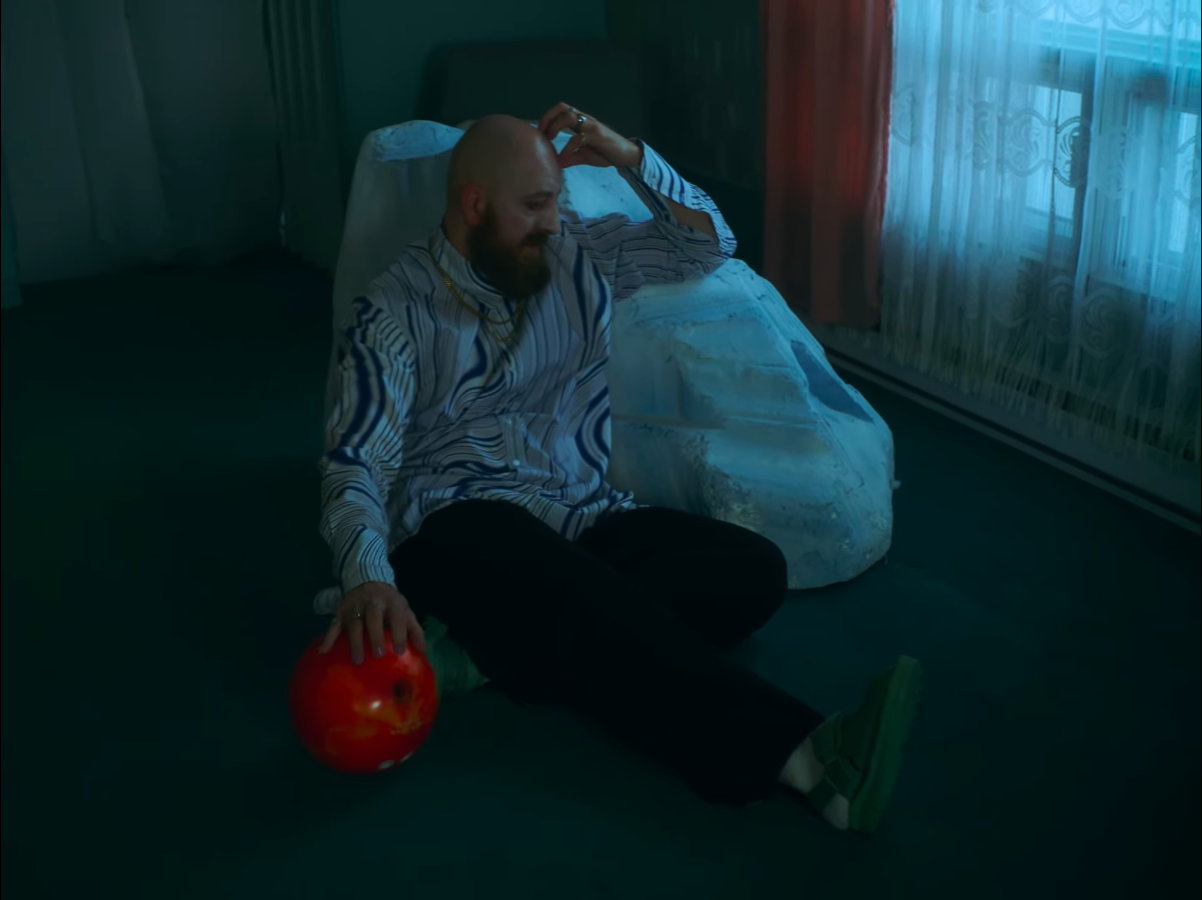 Un homme assis par terre dans une pièce sombre, tenant une pomme rouge, avec une tête rasée et une barbe, portant une chemise à rayures et un pantalon noir, près d'une fenêtre avec des rideaux et un lit en arrière-plan.