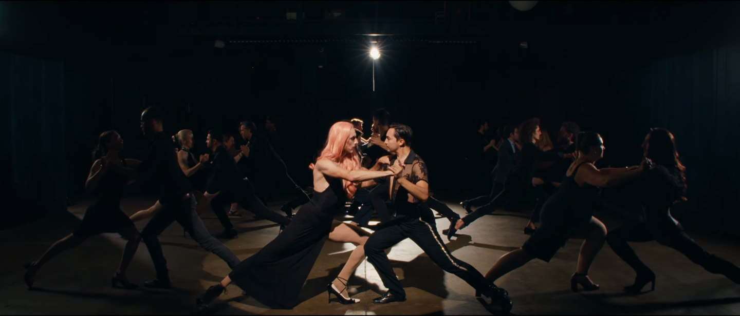 Groupe de danseurs en costume noir se tenant par la main, formant une ligne, dans une pièce sombre avec un éclairage central.
