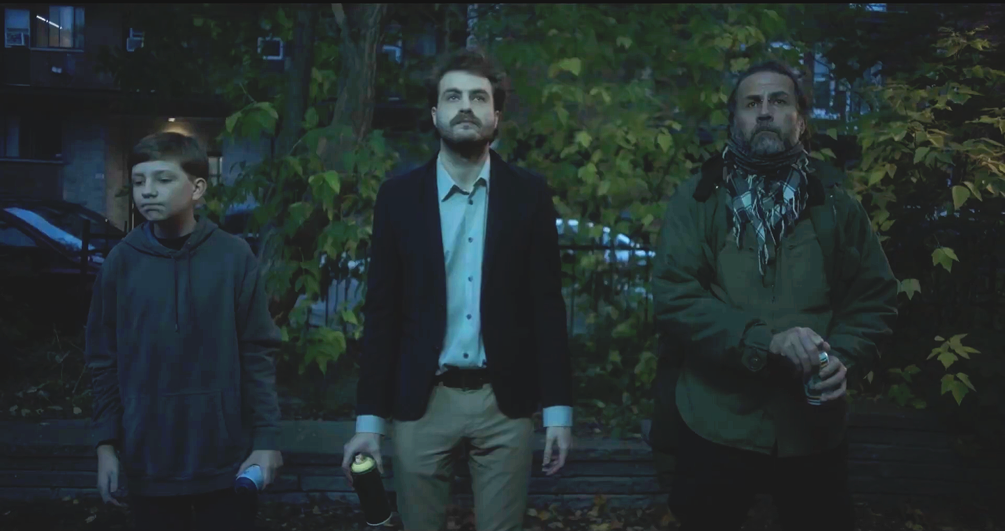 Trois personnes debout face à un arrière-plan de feuillage en soirée, ambiance sombre, beaucoup de feuilles mortes au sol.