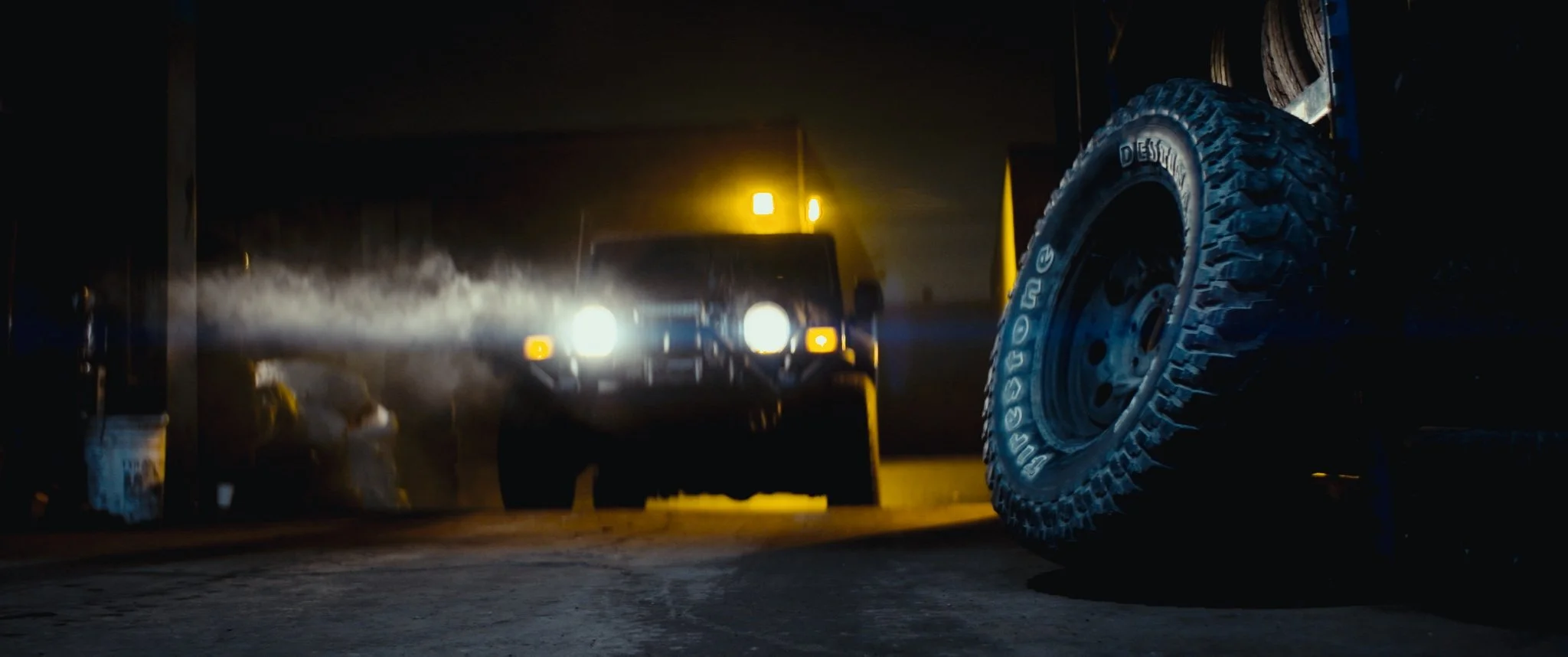 Un voiture noire avec ses phares allumés circule dans une zone sombre, avec une grande roue tout-terrain à proximité à droite et une lumière jaune à l'arrière-plan.