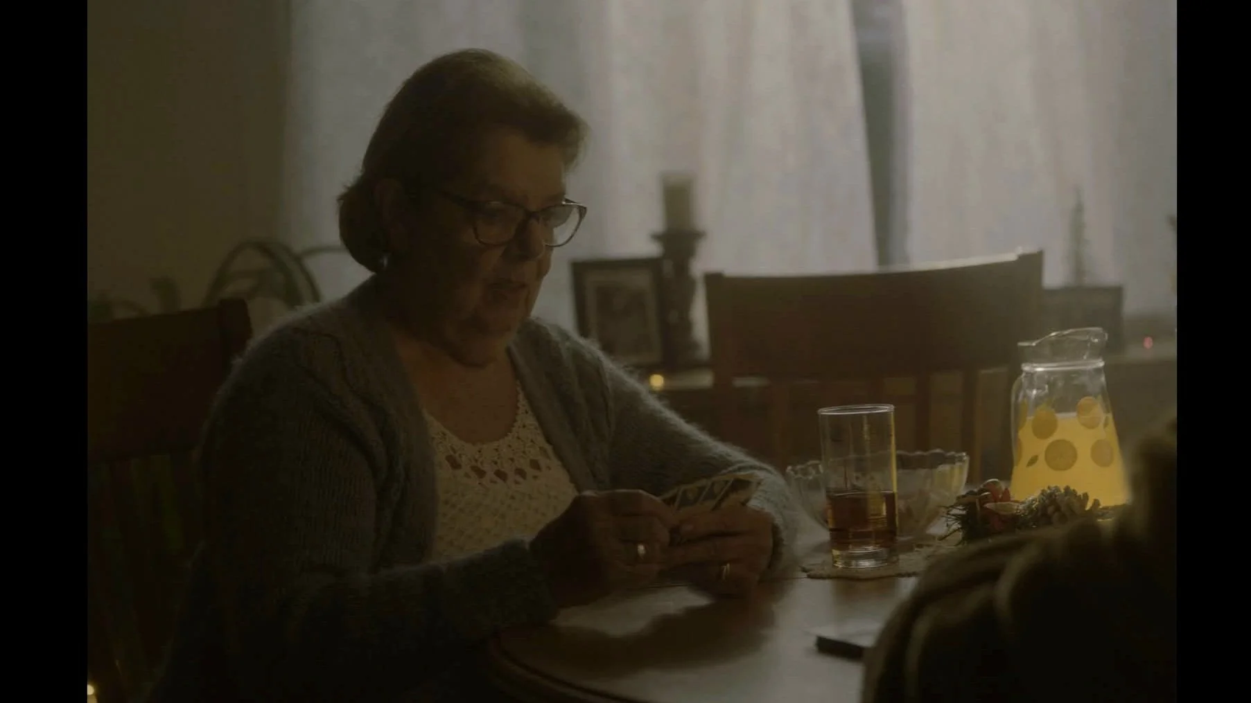 Une femme âgée avec des lunettes, assise à une table, regarde son téléphone. Sur la table, il y a un pichet de boisson citronnée, un verre, un bol, et un verre d'iced tea. La pièce est éclairée doucement avec des rideaux blancs en arrière-plan.