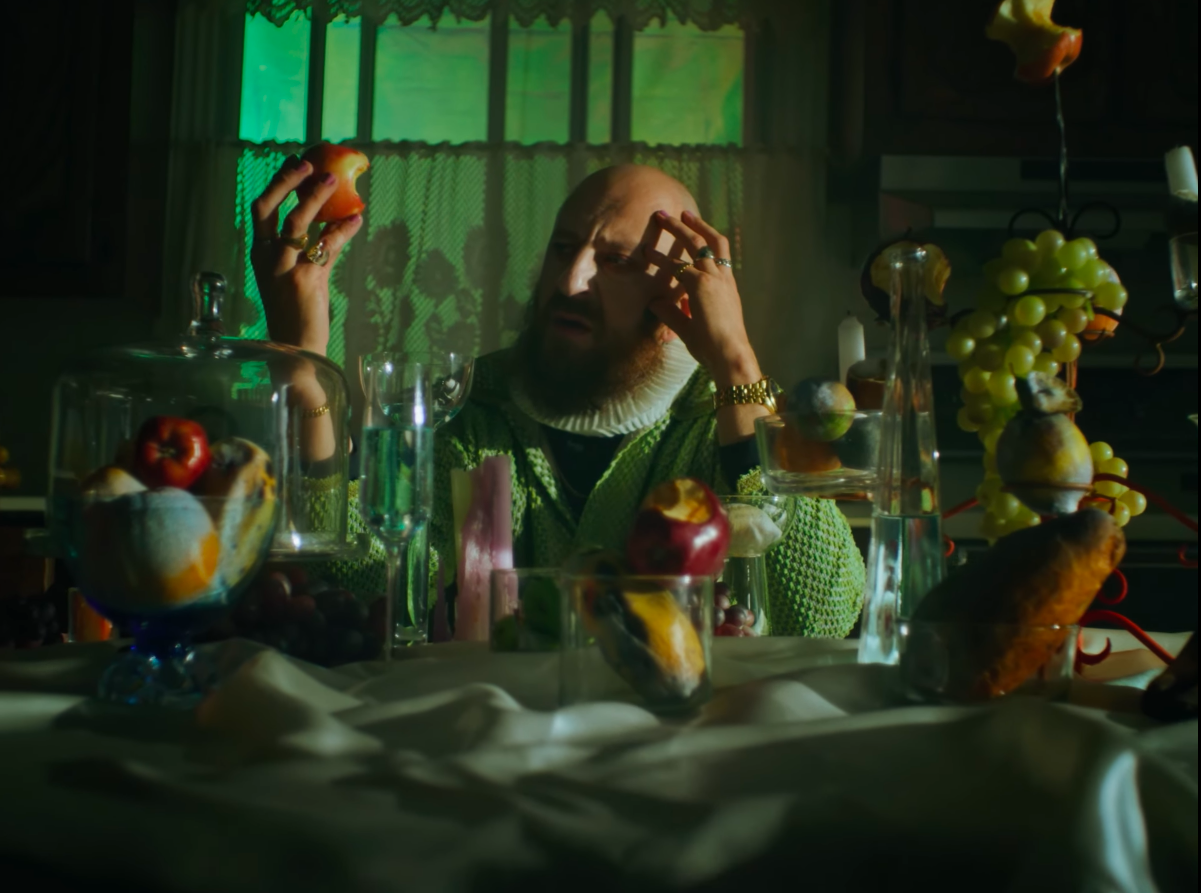 Un homme devant une table remplie de fruits, d'objets et de verre, tenant une pomme dans une main et une autre pomme dans l'autre, dans une pièce sombre avec une lumière verte en arrière-plan.