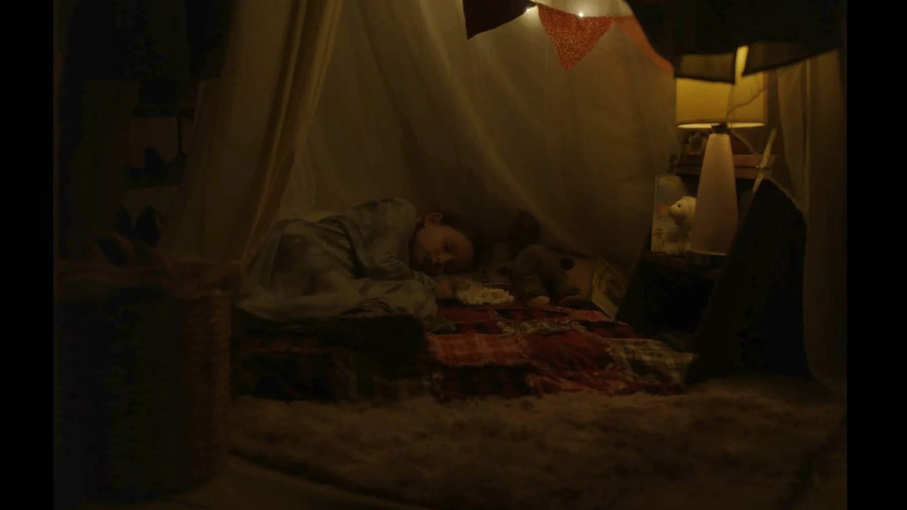 Un enfant qui dort dans un lit dans une tente ou un coin de camping, entouré de plusieurs couvertures et jouets, avec une petite lampe de chevet à côté.