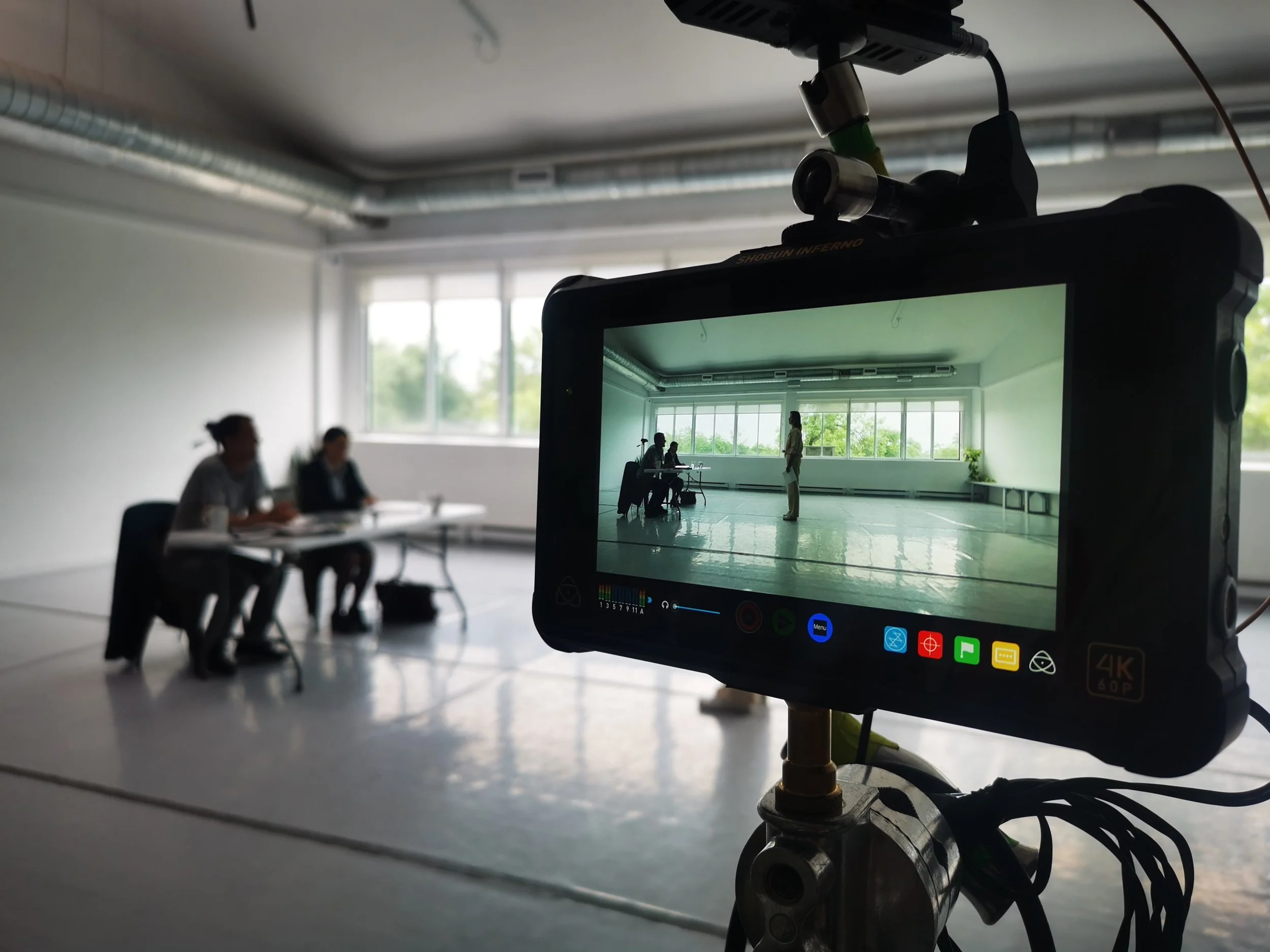 Un écran de caméra de cinéma montrant une scène avec plusieurs personnes dans une pièce lumineuse, tandis qu'une caméra filme la scène. L'image est prise dans un studio ou une salle de réunion.