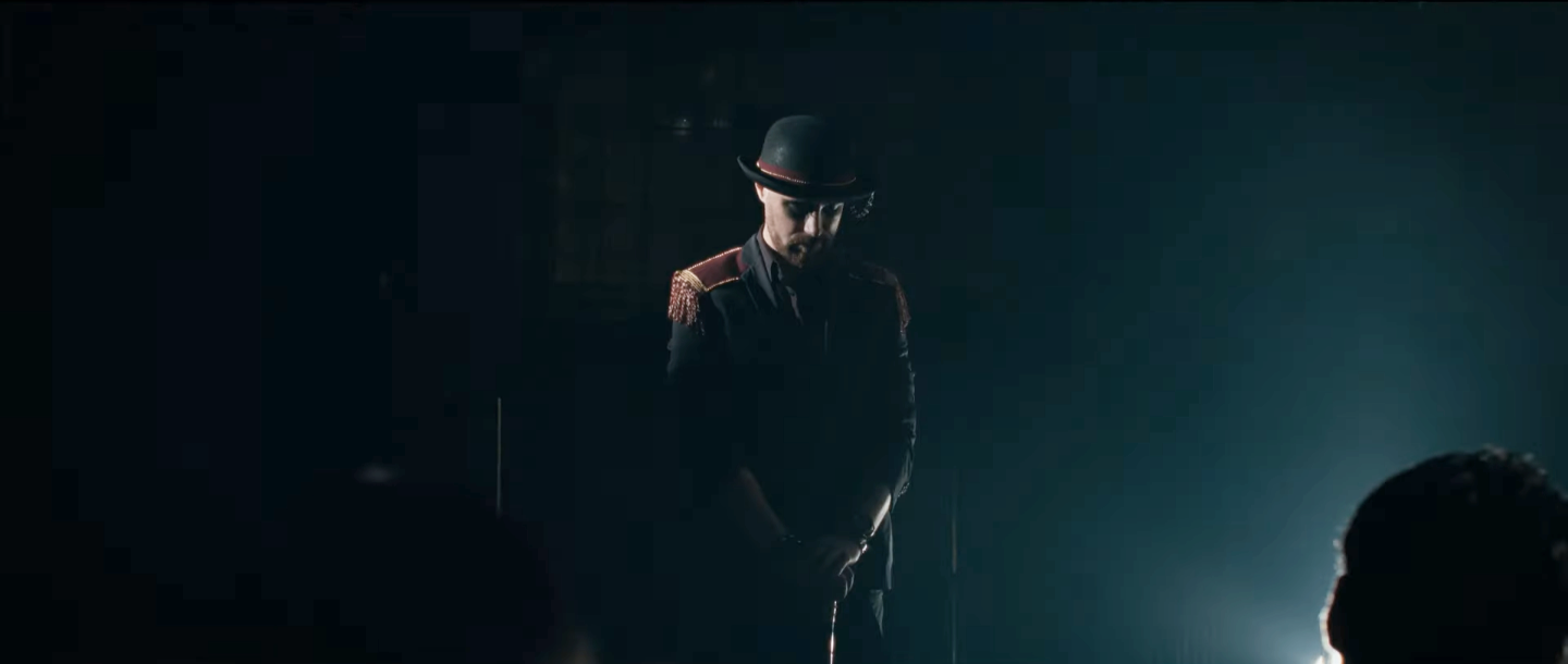 Un homme déguisé en militaire ou en officier, portant un chapeau et une veste avec des épaulettes, dans une ambiance sombre avec une lumière qui illumine partiellement son visage.