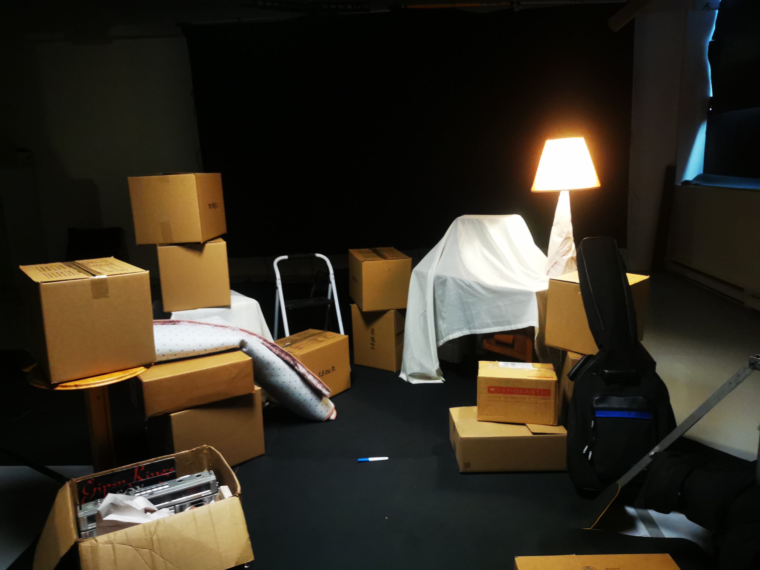 Une pièce sombre avec plusieurs cartons en carton dispersés, une lampe allumée, une chaise, une couverture, et des objets divers sur une table. Il semble s'agir d'un espace de stockage ou de déménagement.