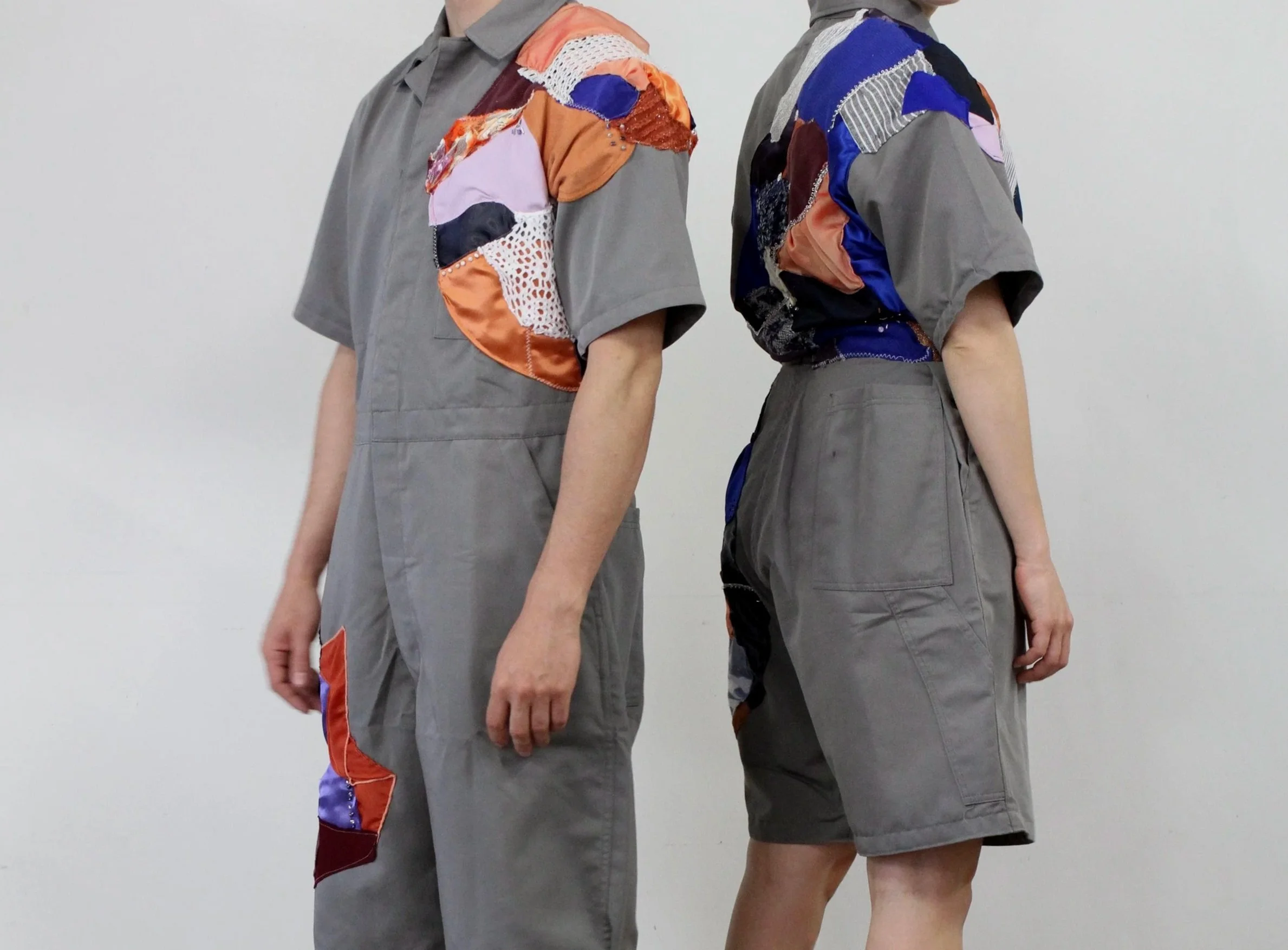Deux personnes debout côte à côte, portant des vêtements gris avec des détails en patchwork colorés sur les épaules et les manches, contre un fond blanc.