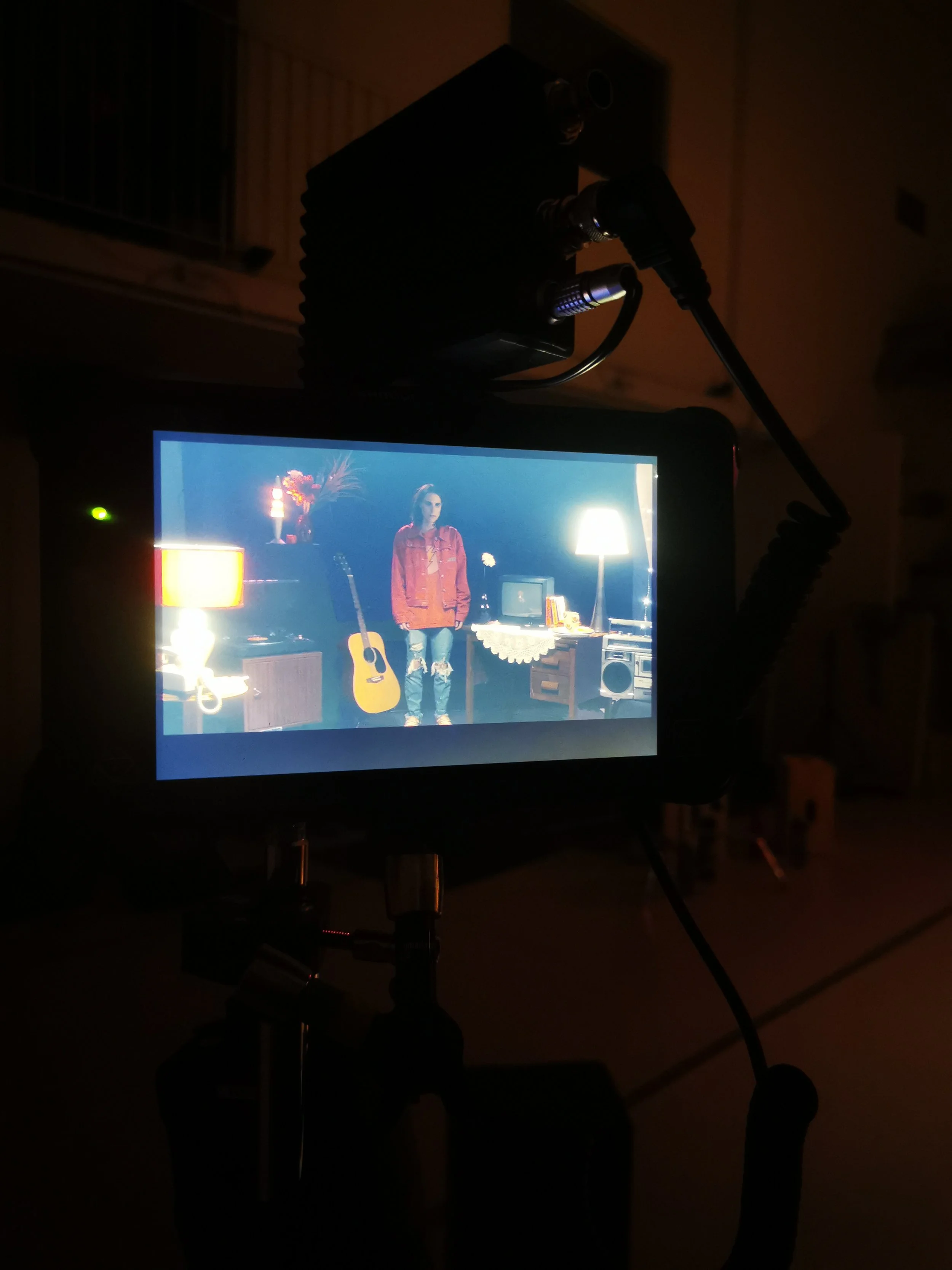 Une caméra de film en prise de vue dans une pièce sombre, affichant une scène avec une femme en rouge devant des meubles et une guitare.