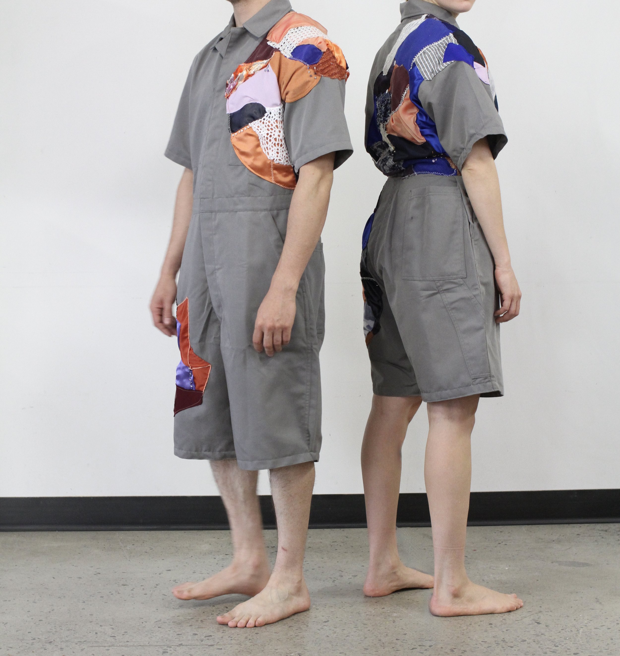 Deux personnes debout dos à dos, portant des vêtements gris avec des détails colorés. La personne à gauche porte une chemise à manches courtes et un pantalon, l'autre porte une robe ou une jupe. Elles se tiennent sur un sol en béton, contre un mur bl