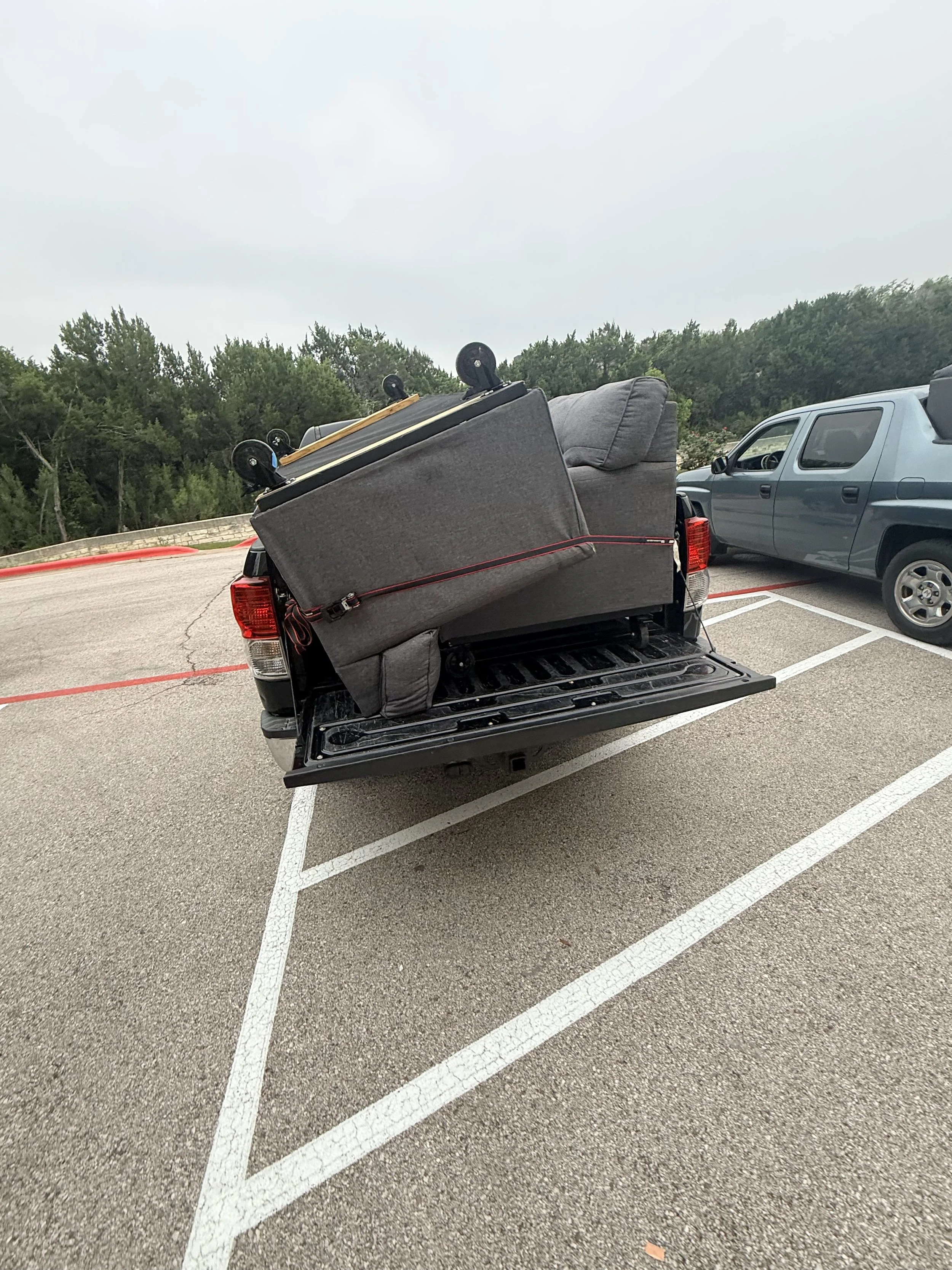 Back of a pickup truck carrying a sofa and an overturned skateboard in a parking lot with other cars and greenery in the background.
