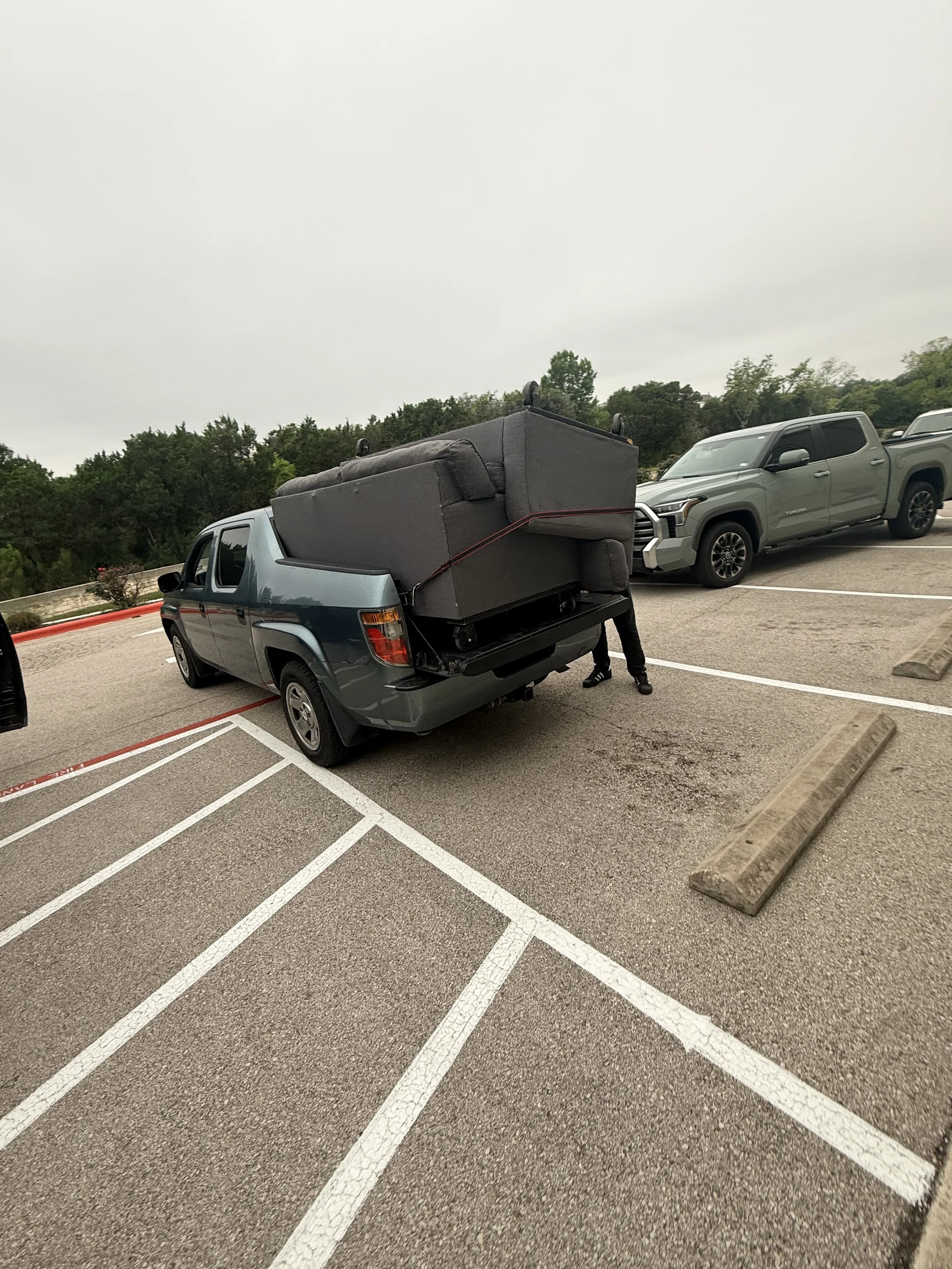 A parking lot with empty spaces, a pickup truck, and an SUV. A person is loading a large, dark-colored sofa onto the back of the pickup truck. The sky is overcast and there are trees in the background.