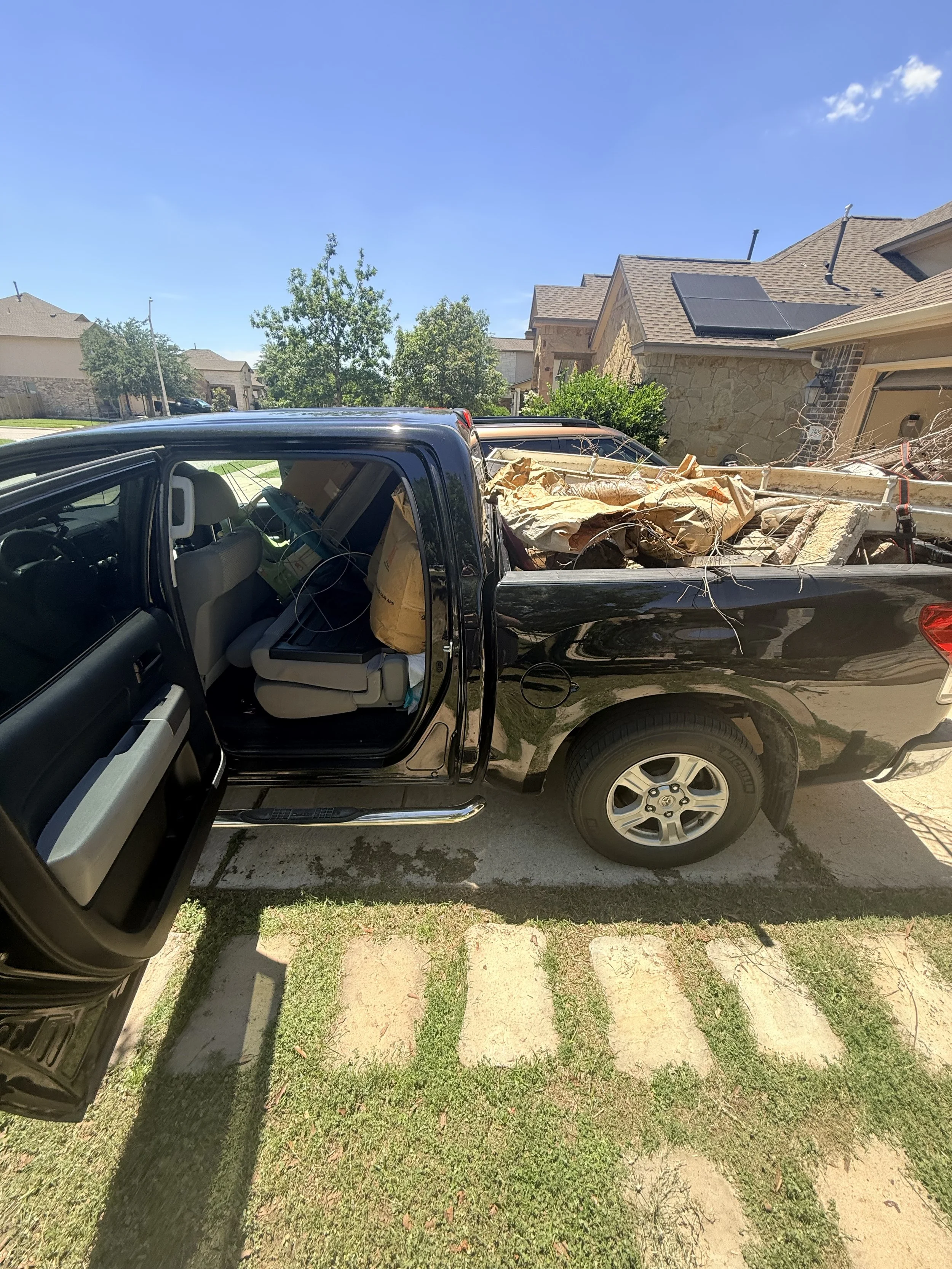 A black pickup truck parked on a partial concrete and grass driveway with a pile of construction debris in the truck bed. In the background, there are houses with trees and a blue sky.