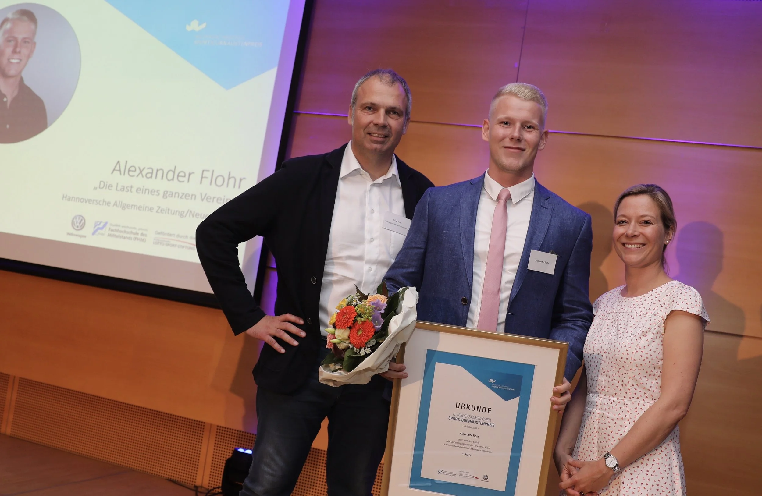 Drei Menschen bei einer Preisverleihung stehen vor einer Wand. Einer hält einen großen Rahmen mit Urkunde, eine Person hält einen Blumenstrauß. Im Hintergrund ist ein Bildschirm mit Text und einem Foto einer Person.
