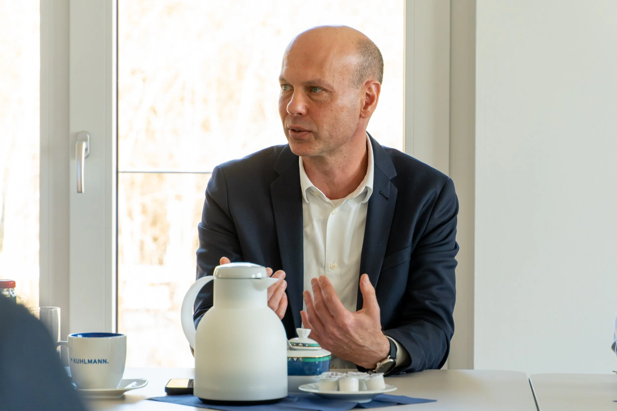Mann sitzt an einem Tisch hinter einer großen Fensterscheibe, spricht und gestikuliert mit den Händen. Auf dem Tisch stehen eine weiße Kaffeekanne, eine Tasse mit dem Schriftzug "KUHLMANN", eine unterste Platte mit kleinen Schälchen und eine Kaffeemaschine.