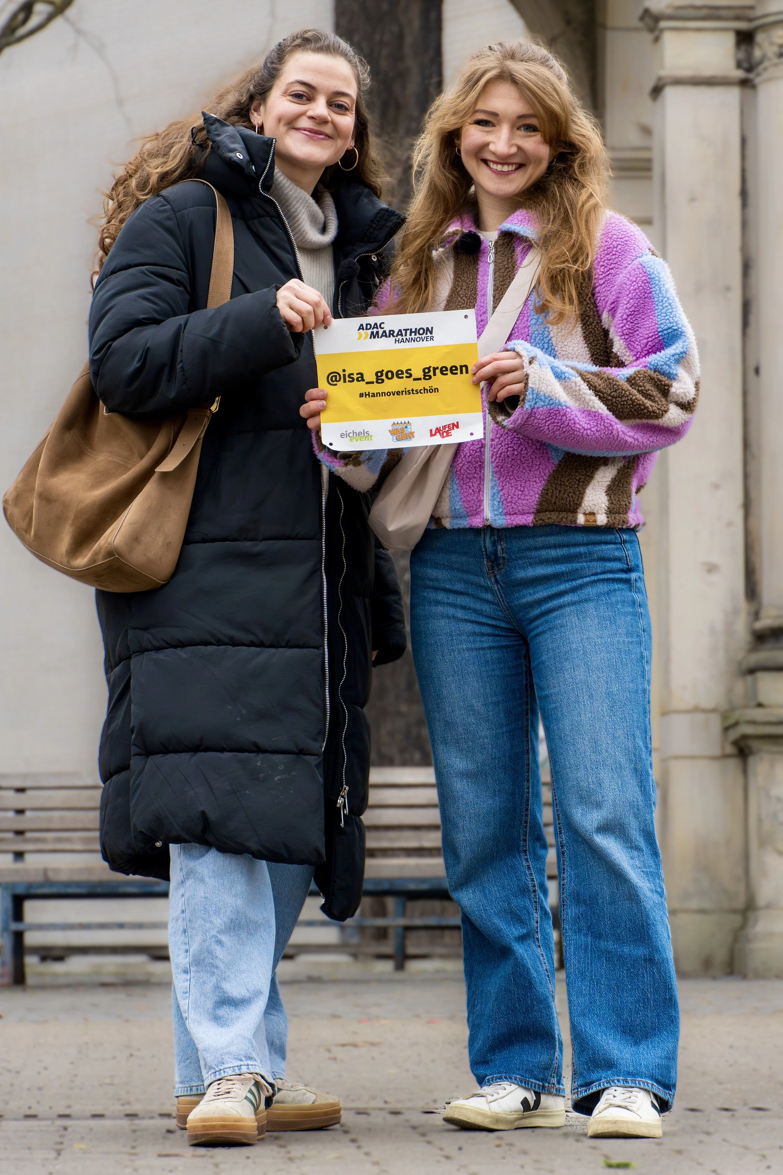 Zwei junge Frauen stehen nebeneinander vor einem Gebäude, eine hält ein Schild mit dem Instagram-Handle @isa_goes_green, beide lächeln. Es ist kalt und sie tragen warme Kleidung.