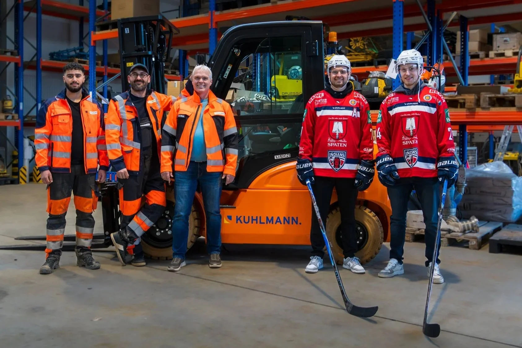 Sechs Männer stehen vor einem orangefarbenen Kuhlmann-Gabelstapler in einem Lagerraum. Vier Männer tragen Eishockeytrikots, zwei Männer tragen orange Sicherheitskleidung. Sie sind alle in einer Lagerhalle mit Regalen und Kartons im Hintergrund.