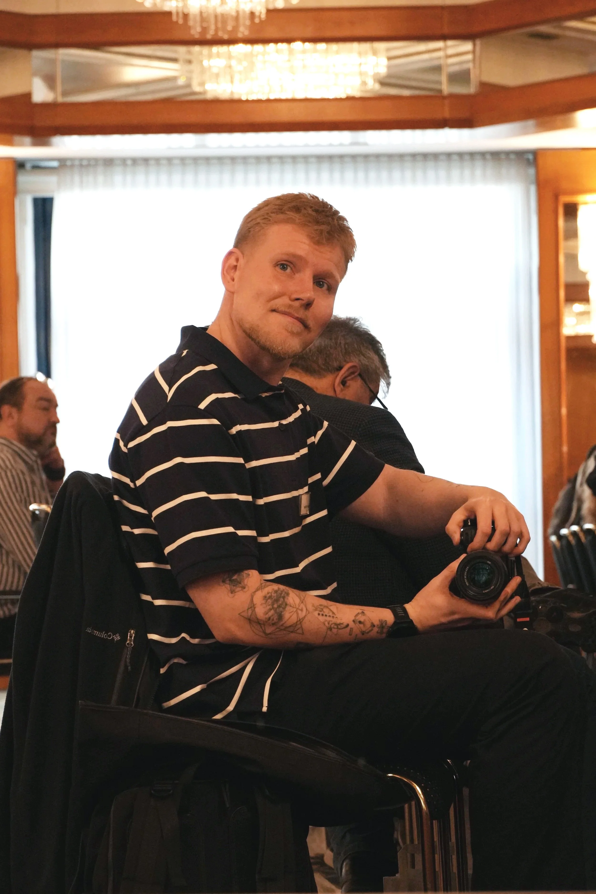 Ein junger Mann mit roten Haaren und Tattoos auf den Armen hält eine Kamera in der Hand. Er trägt ein schwarz-weiß gestreiftes Hemd. Im Hintergrund sind andere Menschen zu sehen, zudem eine helle Fensterfront und eine dekorative Decke mit Kronleuchtern. Der Raum wirkt elegant eingerichtet.