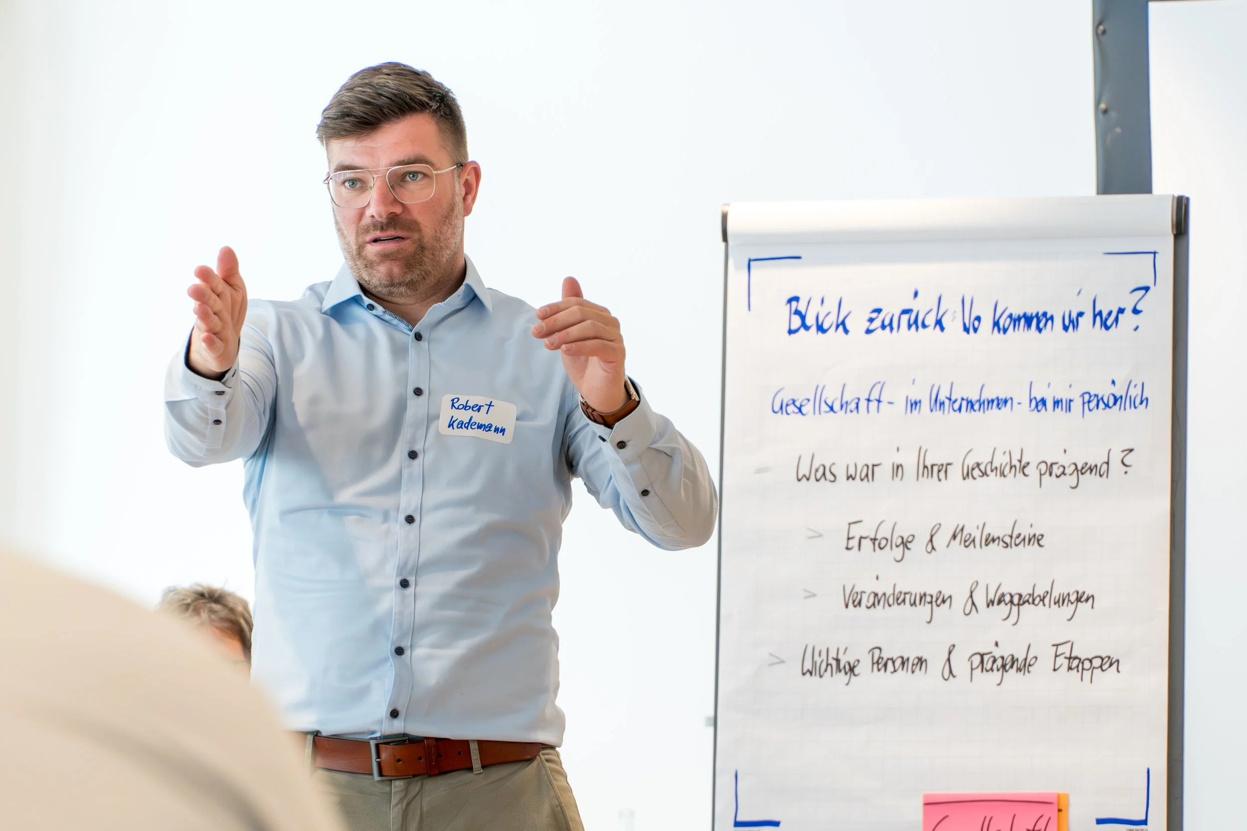 Ein Mann mit Brille und braunen Haaren erklärt etwas vor einer weißen Flipchart, auf der auf Deutsch geschrieben steht. Er trägt ein hellblaues Hemd und eine braune Gürtelschnalle.