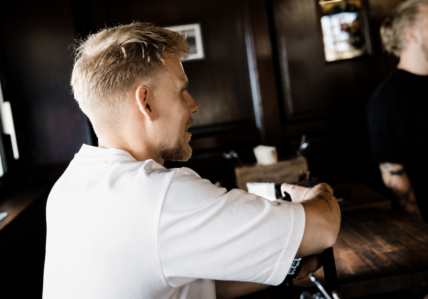 Junger Mann mit blondem Haar, weißen T-Shirt, tätowiert, sitzt an einem Holztisch in einer dunklen Holzraumumgebung, blickt nach rechts.