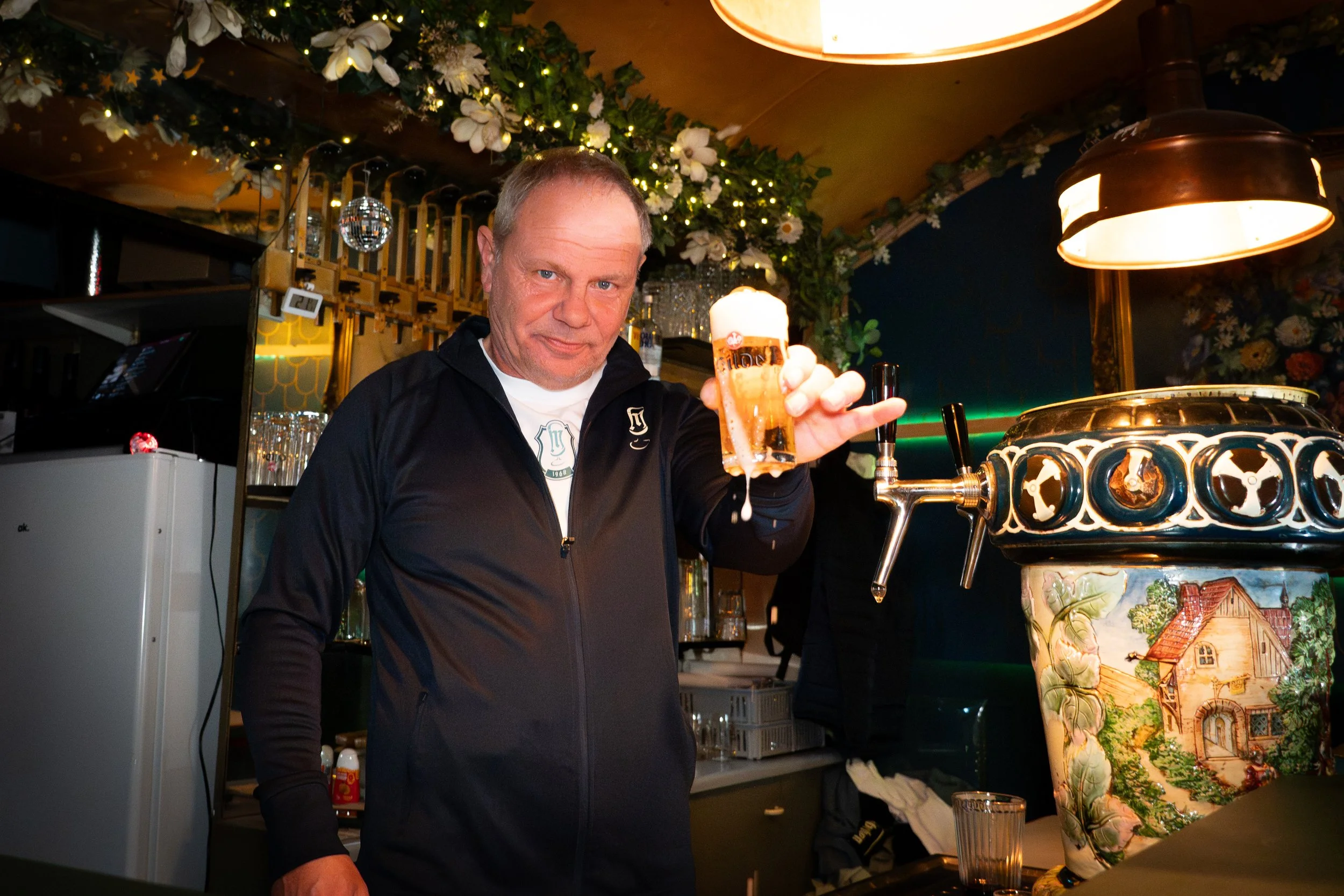 Ein Mann hält ein volles Bierglas in einer Bar mit dekorativen Blumen und Lichterketten im Hintergrund.
