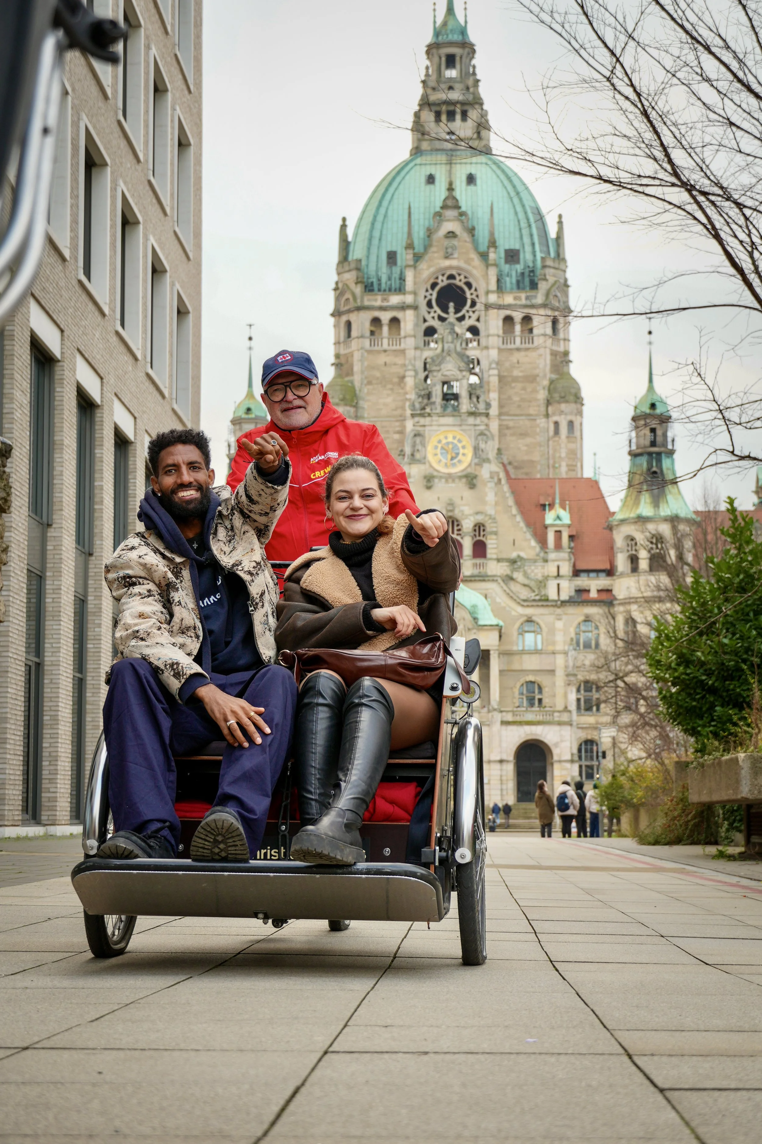 Drei Menschen in einem Rollstuhl auf einer Stadtstraße, im Hintergrund die Türme einer alten Kathedrale, umgeben von Bäumen ohne Blätter, es ist bewölkter Himmel.