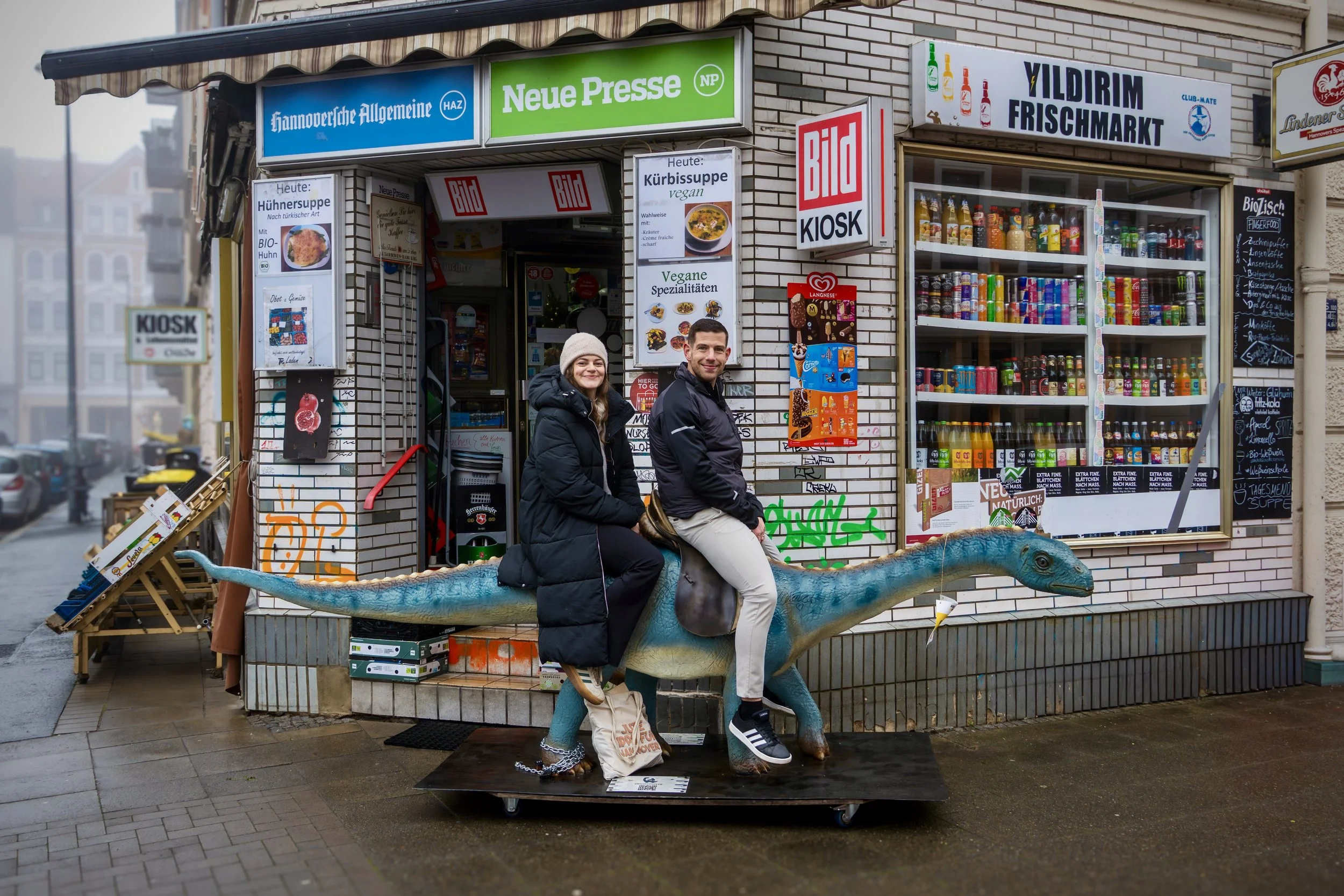Zwei Menschen sitzen auf einem Dinosaurier-Figur vor einem Kiosk, der Restaurants und einen Fischmarkt bewirbt. Das Geschäft befindet sich in einer Stadt, offensichtlich bei regnerischem Wetter.