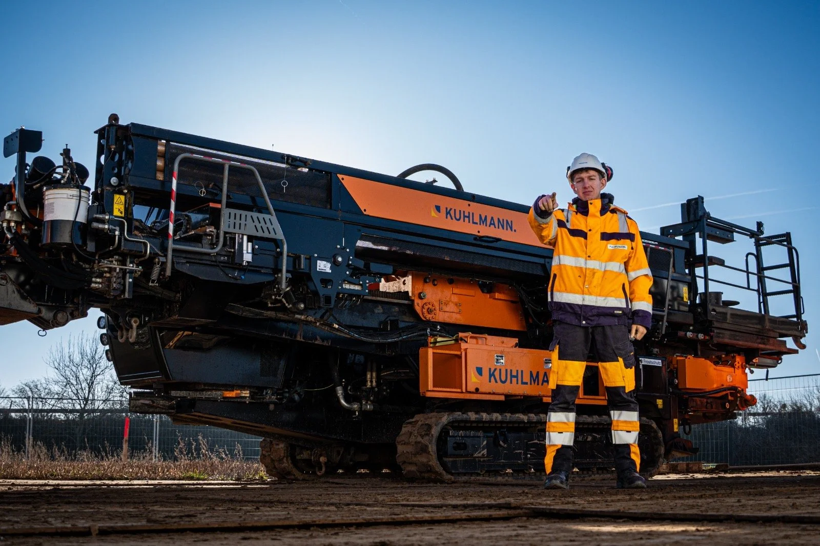 Bauarbeiter in Orange und Schwarz mit Schutzhelm neben großer Baumaschine auf Baustelle.