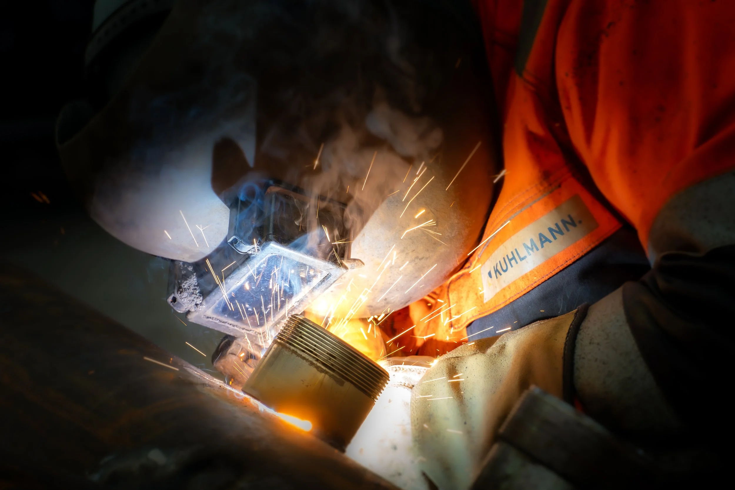 Schweißer arbeitet an Metallrohr, Funkenflüge beim Schweißen sichtbar.