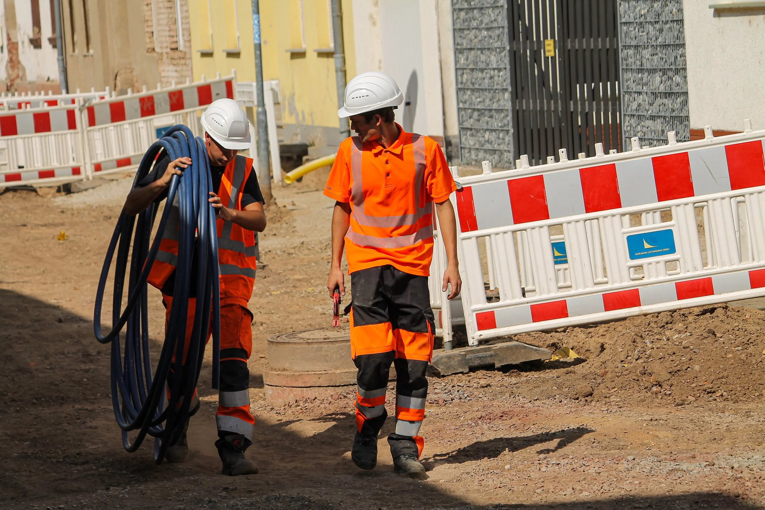 Zwei Bauarbeiter in Sicherheitshelmen und orange Warnschutzwesten auf einer Baustelle, einer trägt Kabel, die andere steht neben einem Mannlochdeckel.