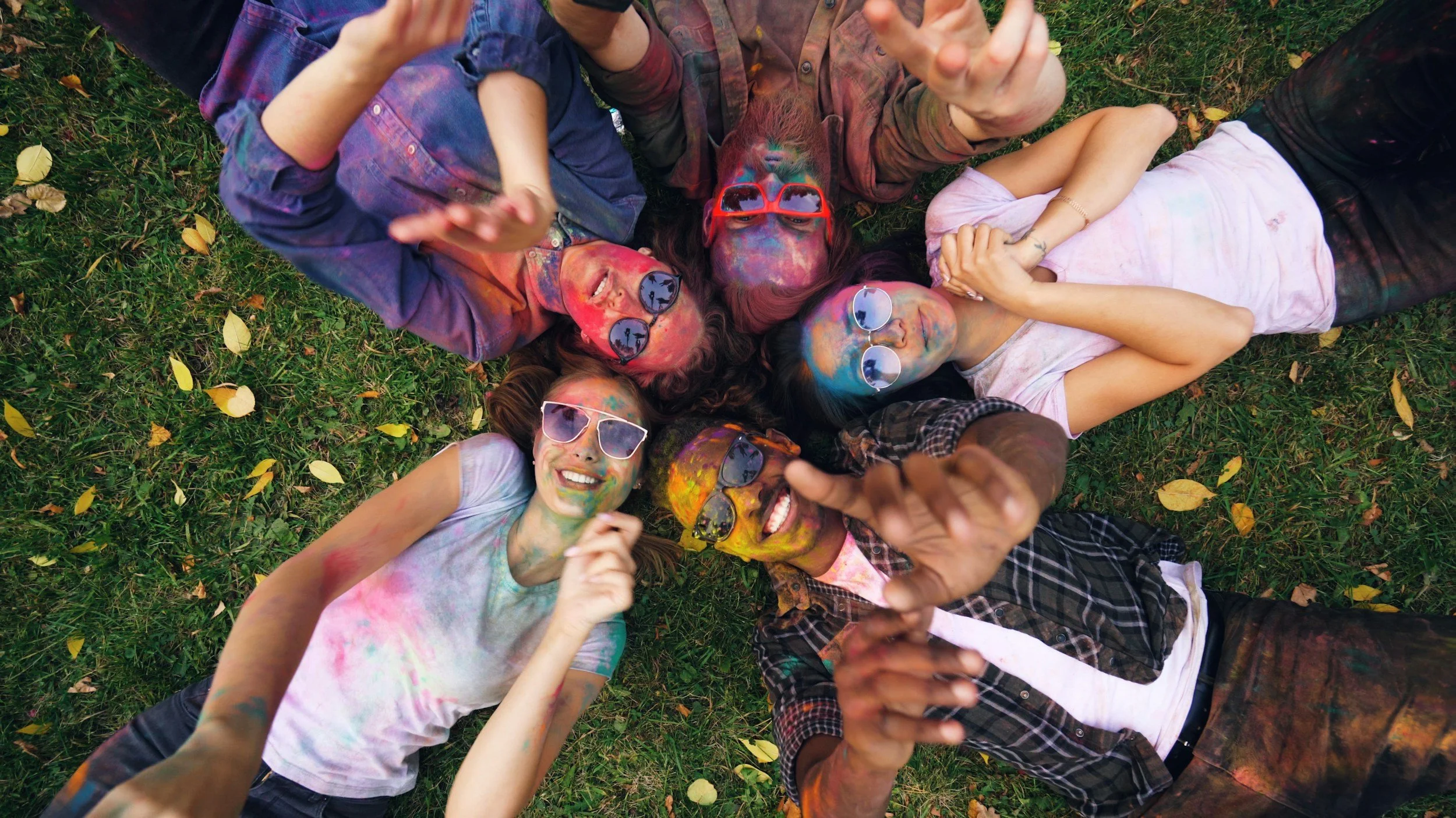 Group of friends lying on grass with colorful powder on their faces and clothes, celebrating Holi, wearing sunglasses, and smiling.