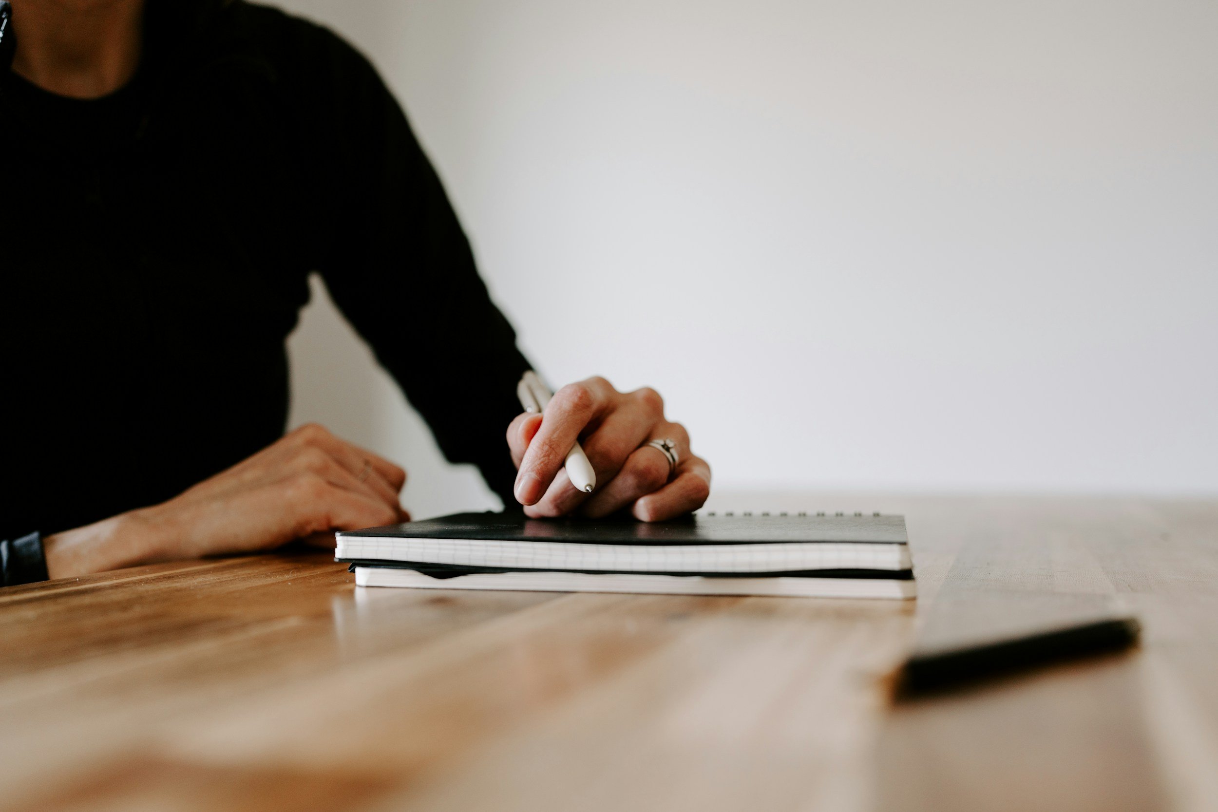 Close-up of a person signing a notebook with a white pen, sitting at a wooden table, with a plain white wall in the background.