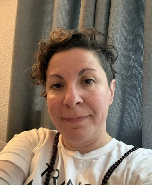 A woman with short, curly hair and a slight smile taking a selfie in front of gray curtains, wearing a white shirt and a black beaded necklace.