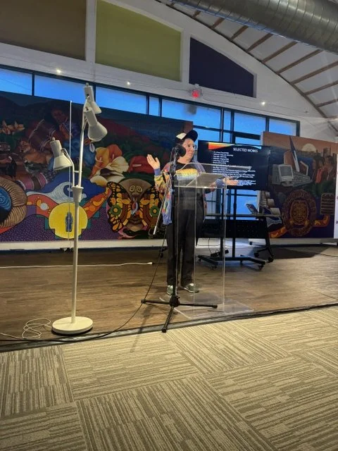 Person speaking at a podium with a microphone, in front of a colorful mural and a large screen displaying a presentation, inside a modern building with high ceilings.