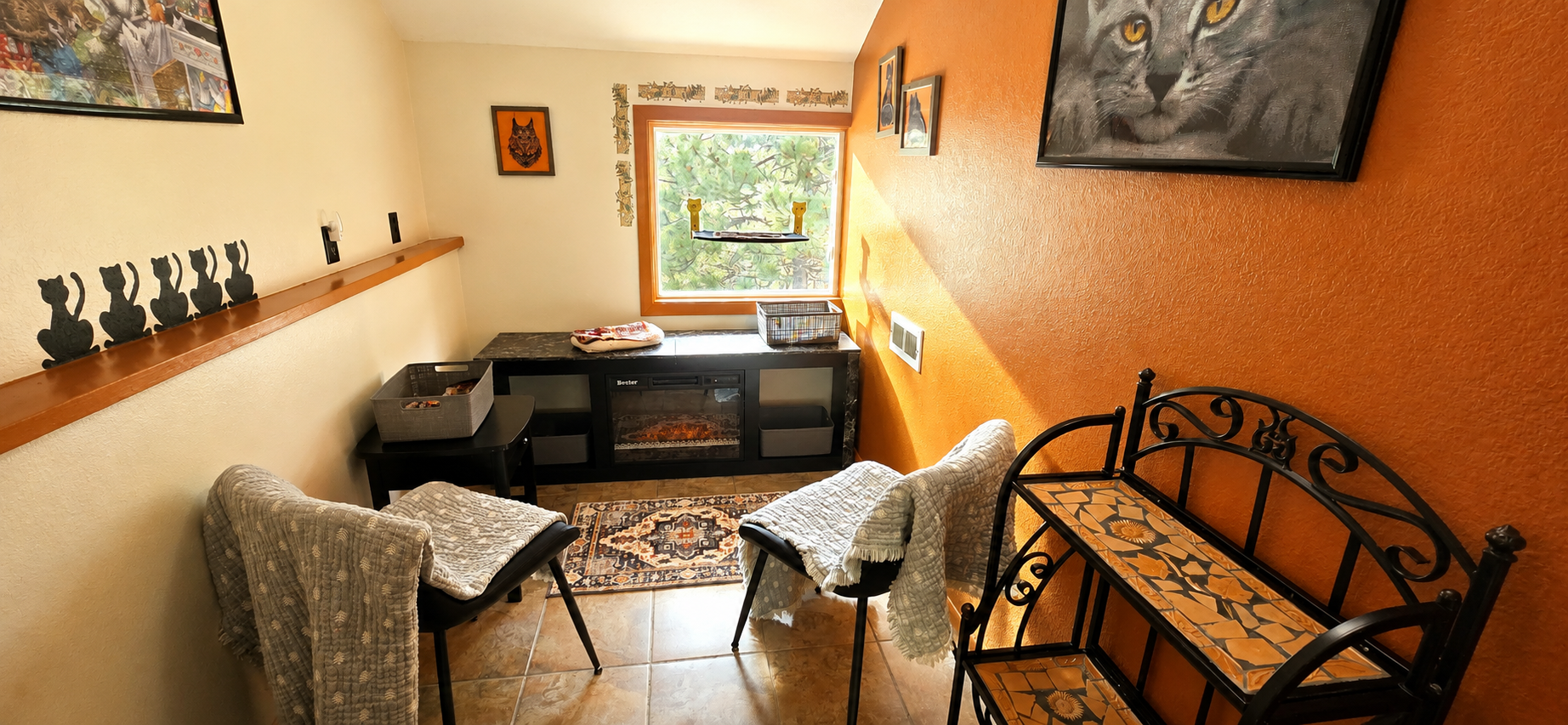 Cozy room with orange and beige walls, decorated with cat-themed artwork, a small electric fireplace, a window, and black metal furniture with floral tiles.