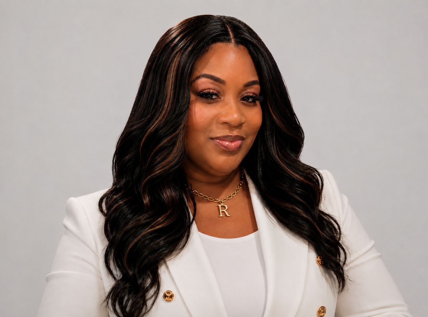 A woman with dark, wavy hair and a white blazer, wearing gold jewelry, and looking at the camera with a slight smile.