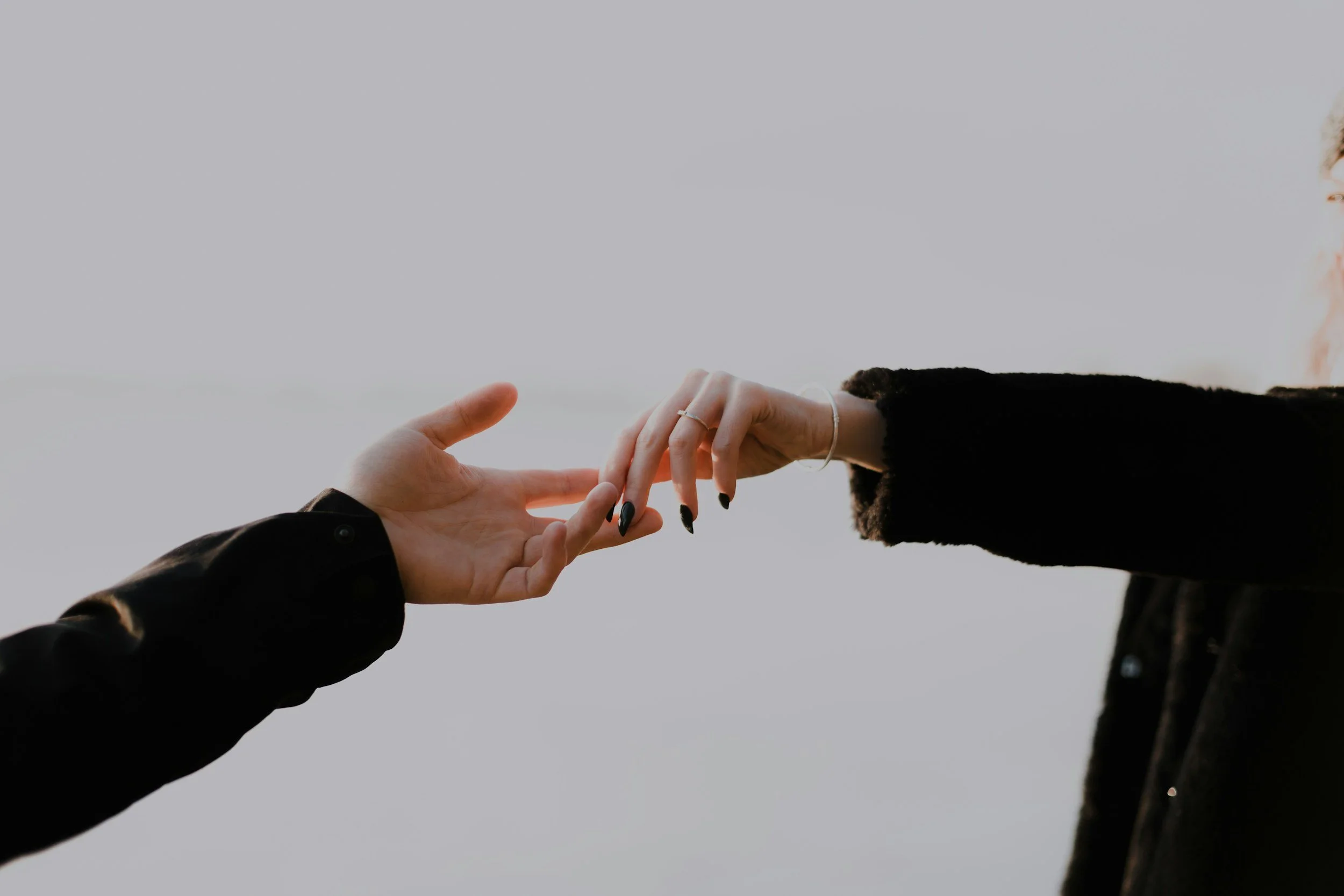 Close-up of two people reaching out to touch each other's hands against a gray sky, one wearing a black jacket and the other with jewelry and a dark sweater.
