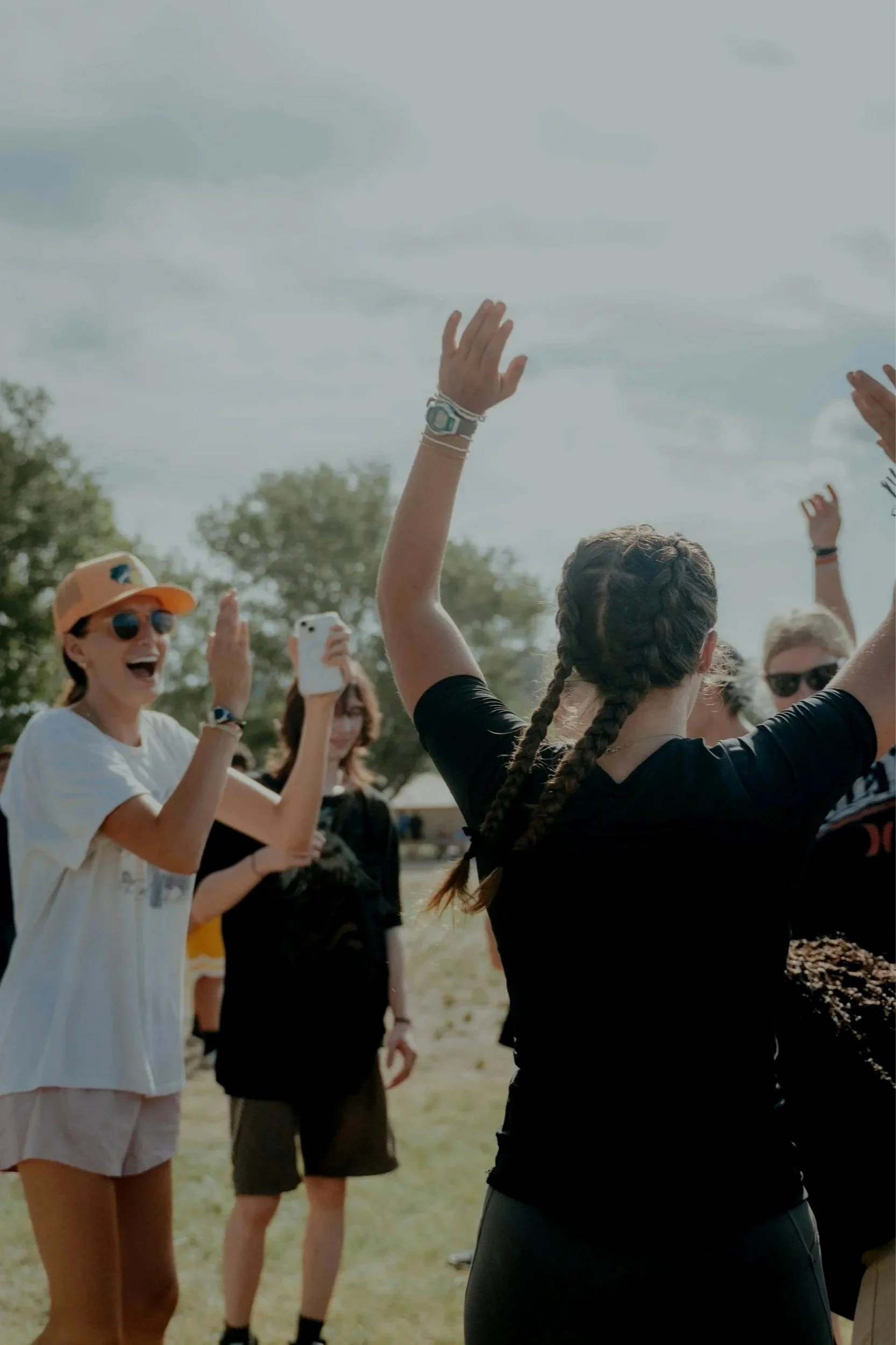 A group of people outdoors, some with their arms raised, talking, taking pictures, and smiling, with trees and a cloudy sky in the background.