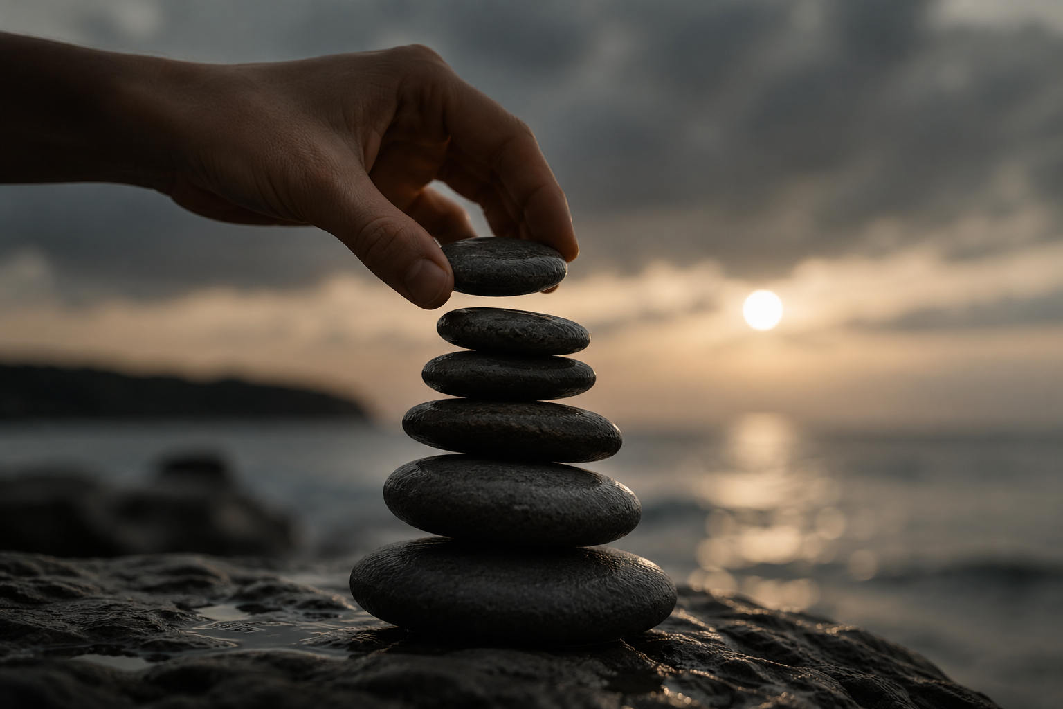A hand stacking black stones on a beach at sunset, with a cloudy sky and the ocean in the background.