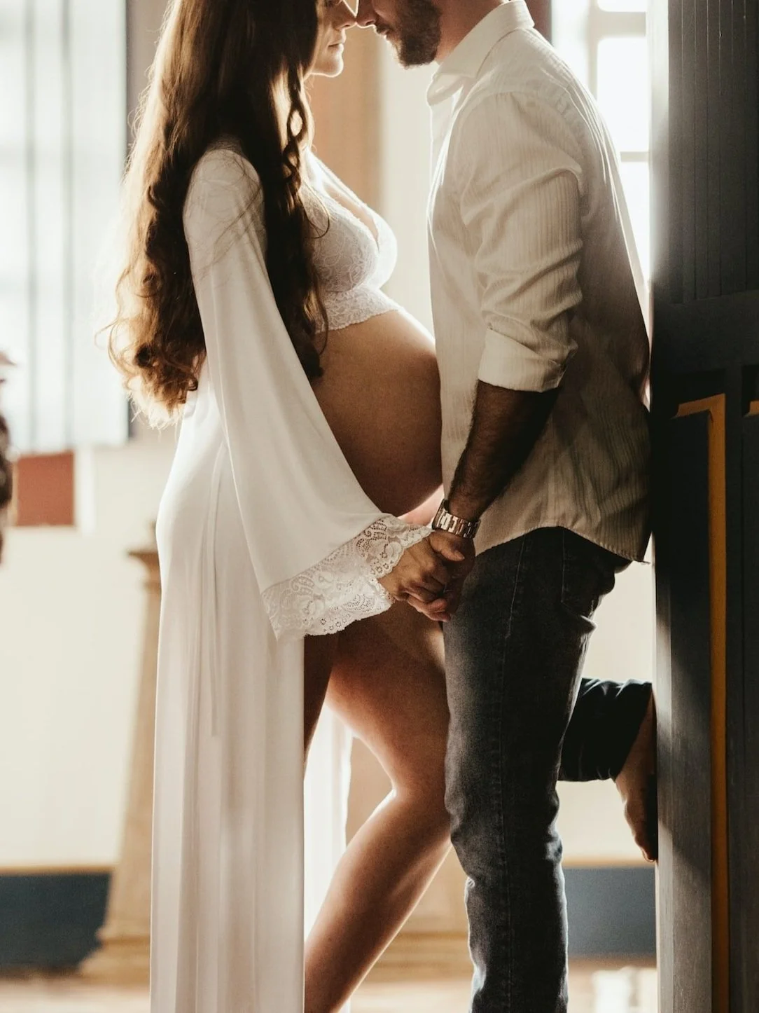 Pregnant woman and man holding hands, standing close indoors, with intimate and affectionate posture.