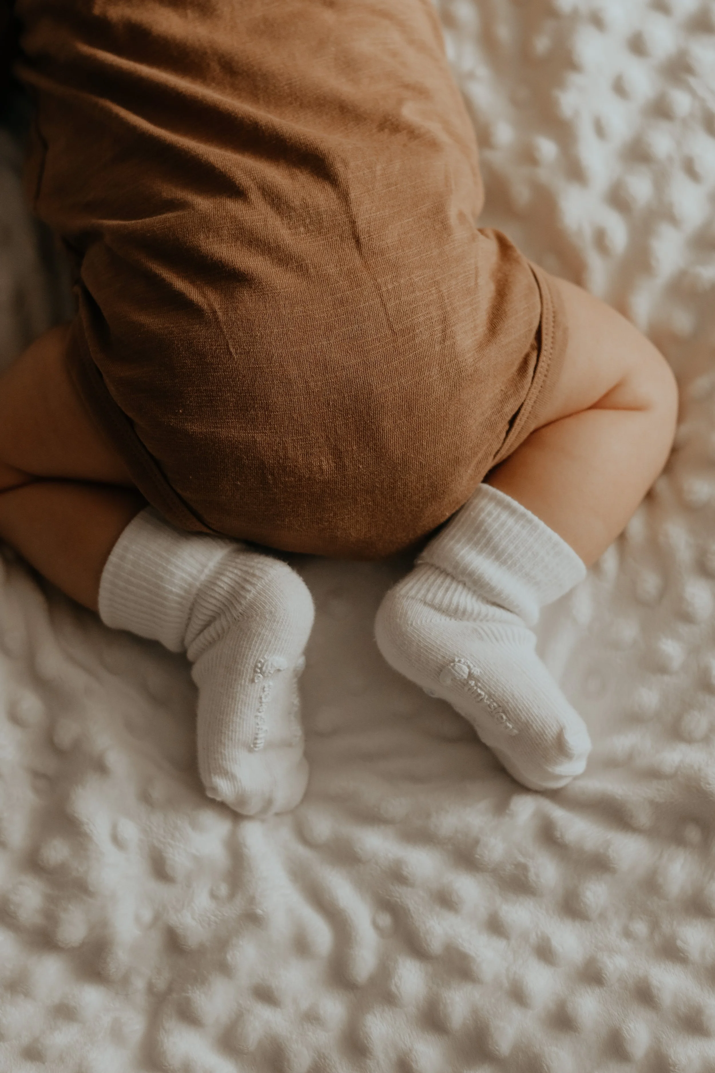 Close-up of a baby crawling on a soft, textured white blanket, wearing tan clothing and white socks.