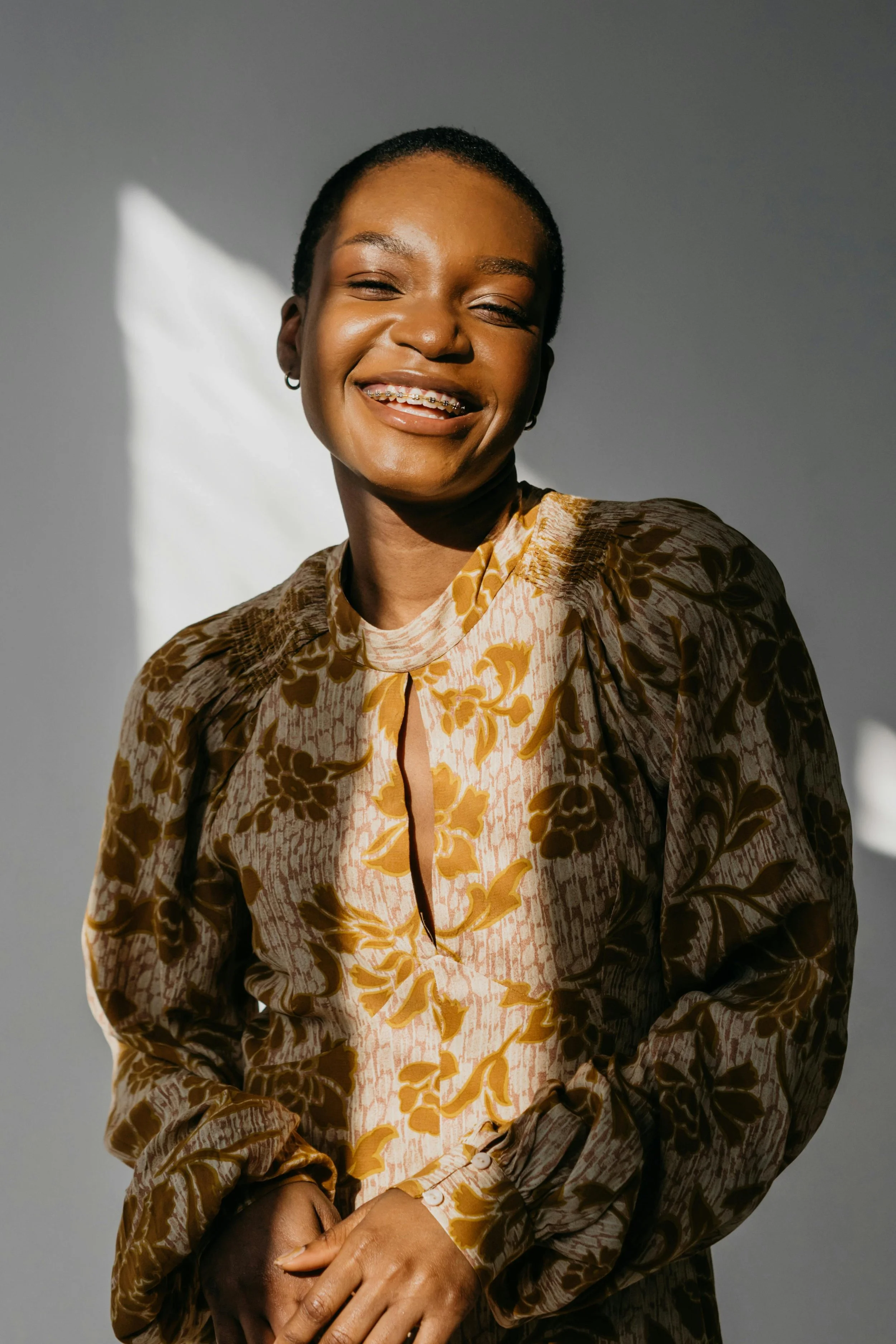 A portrait of a smiling woman with short hair, wearing a patterned dress with yellow and brown floral designs, standing against a gray background with sunlight casting shadows.