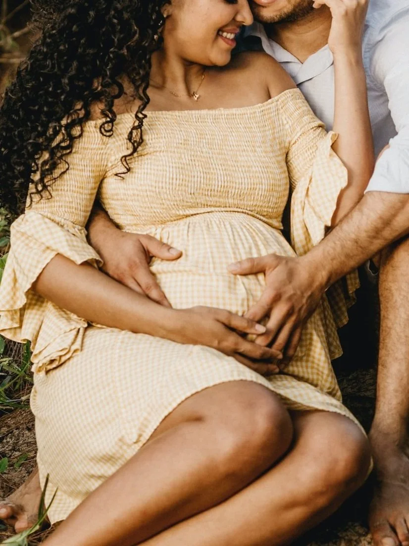A pregnant woman in a yellow checkered dress smiling and gently touching her belly, with a man beside her showing affection, both sitting outdoors.