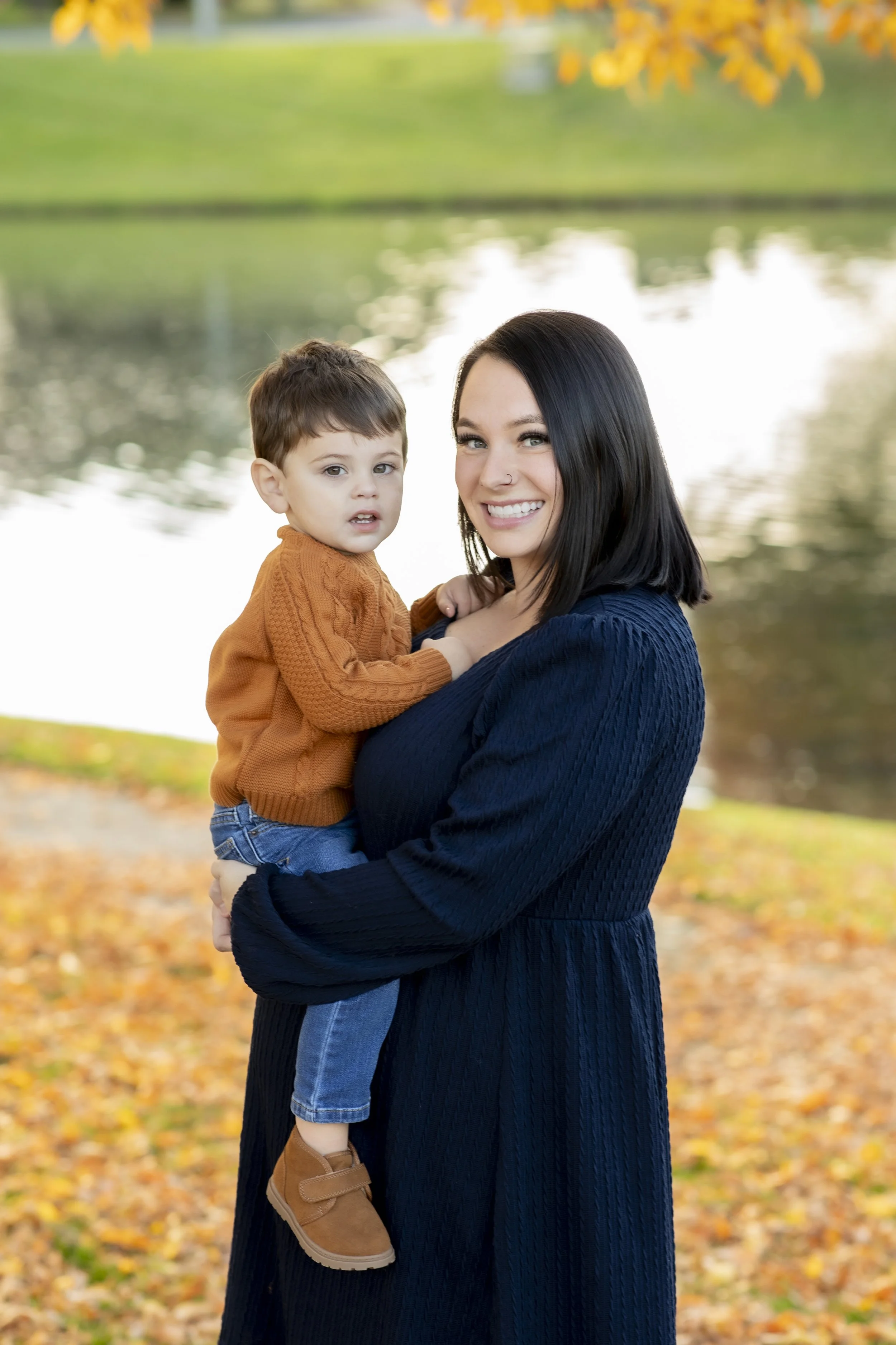A woman with dark hair, wearing a navy blue sweater, holding a young boy with brown hair, wearing an orange sweater, standing outdoors near a body of water with fallen autumn leaves on the ground.