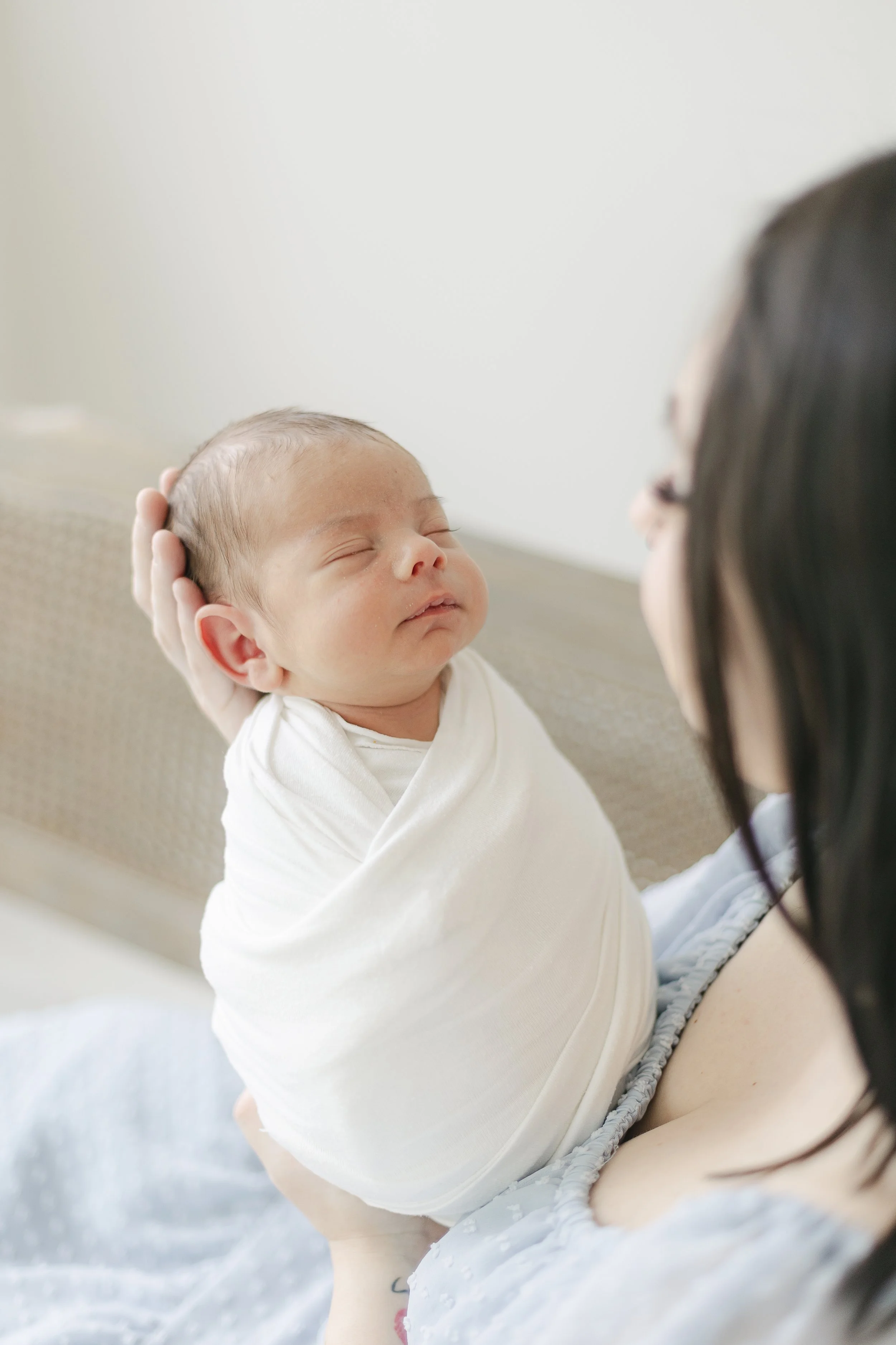 A woman holding a sleeping baby with a content expression, near her face, in a softly lit room.